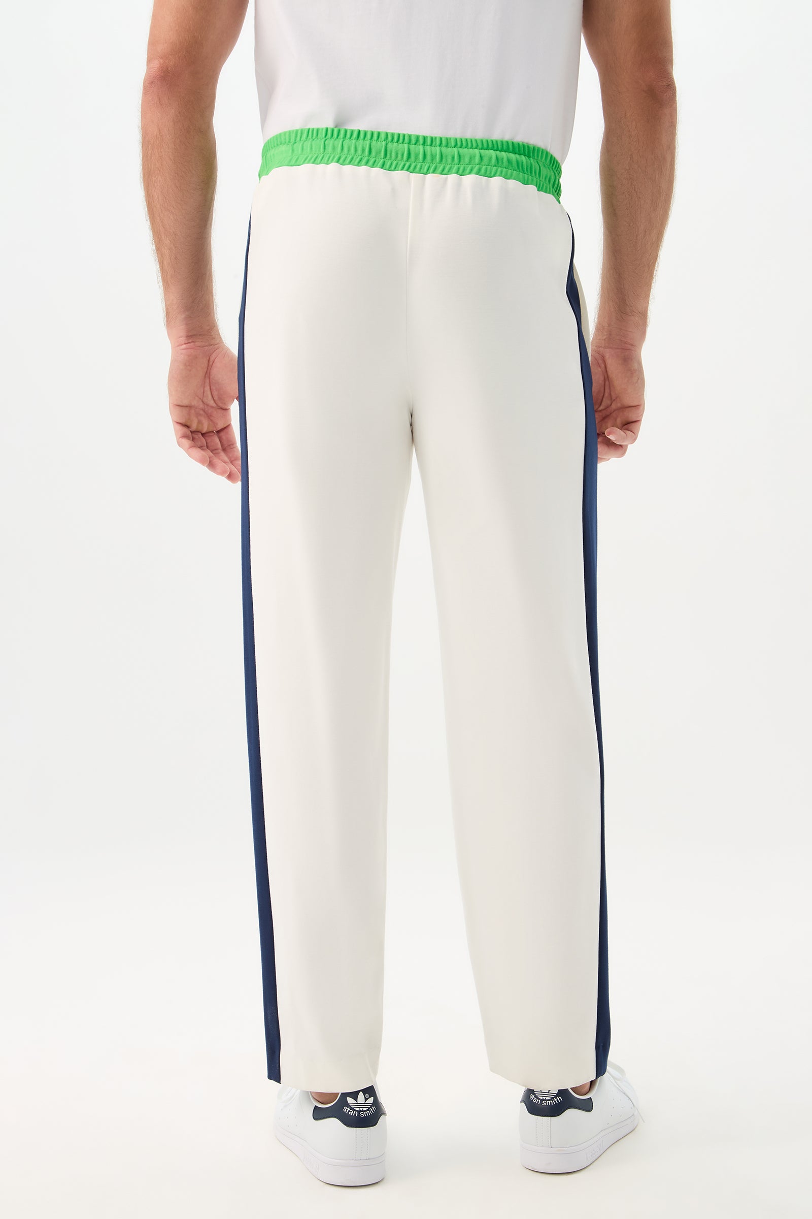A person stands facing away, wearing the Tennis Track Pant - Cozy Ponte with a green elastic waistband, dark blue side stripes, and white Adidas sneakers against a plain white background.