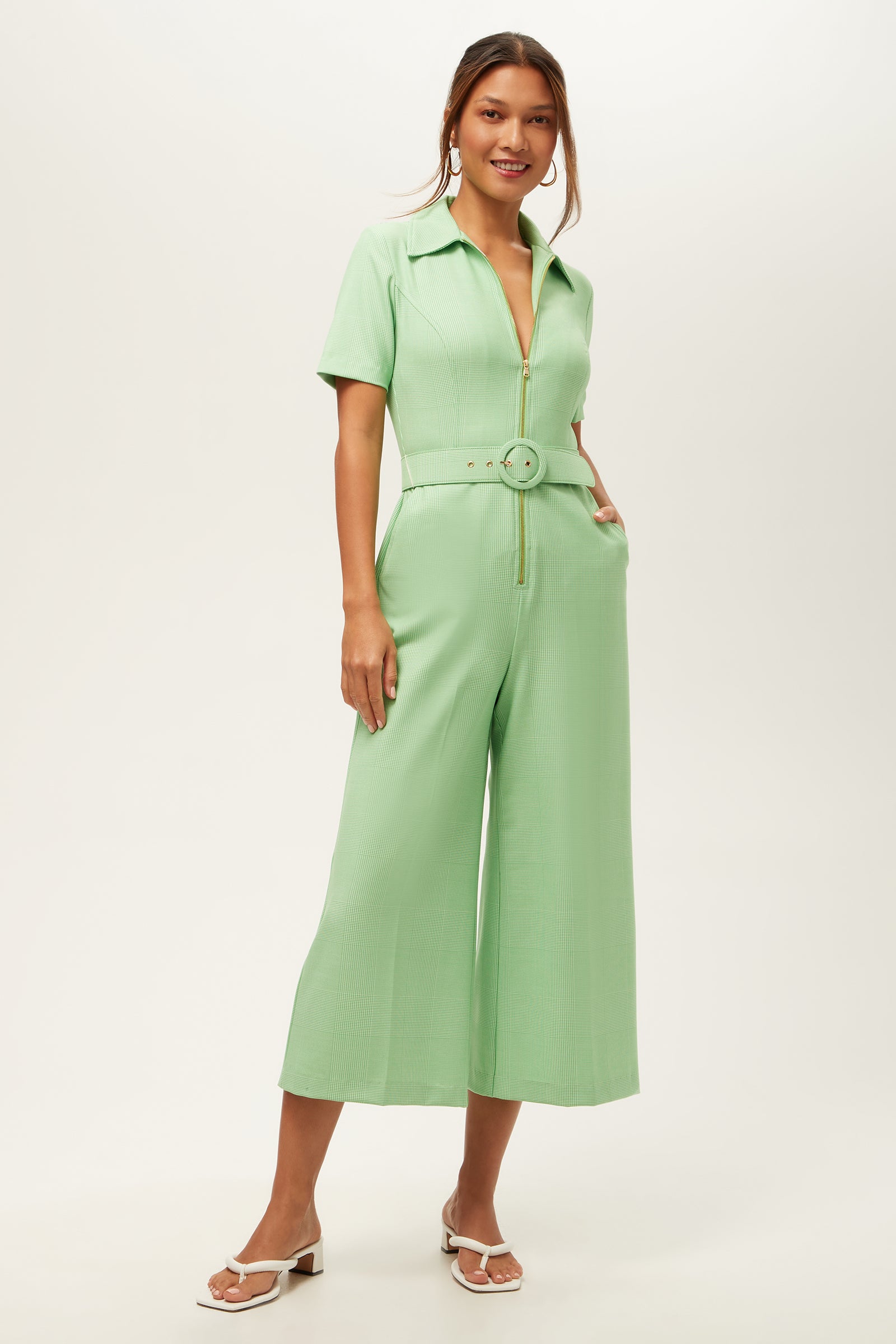 A woman smiles in the Jannise 3 Jumpsuit - Game Time Glen Plaid, featuring a grass court green/whitewash color, collared neckline, wide legs, short sleeves, front zipper, and matching belt. She pairs it with white sandals; her hair is loosely tied back.