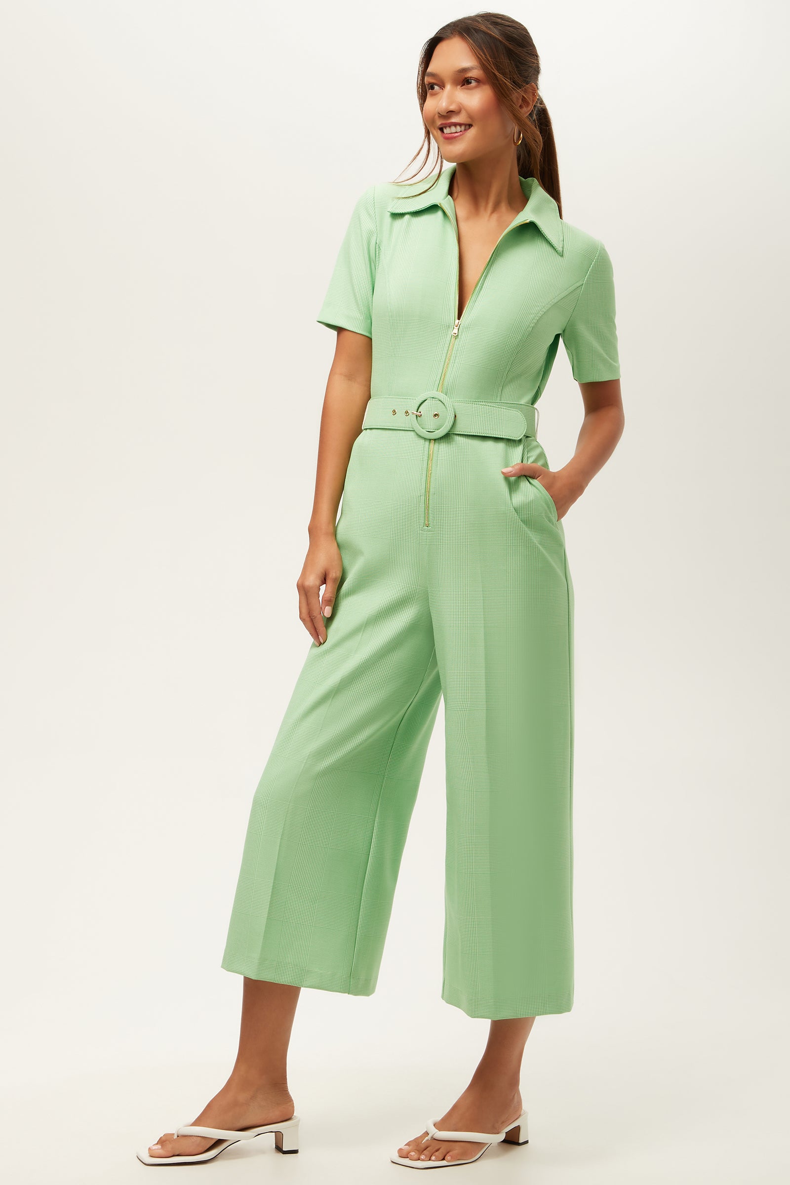 A woman stands smiling, wearing the Jannise 3 Jumpsuit in Game Time Glen Plaid. The jumpsuit features a zip front, short sleeves, wide legs, and a matching belt. Her hair is tied back and she pairs the look with white sandals.