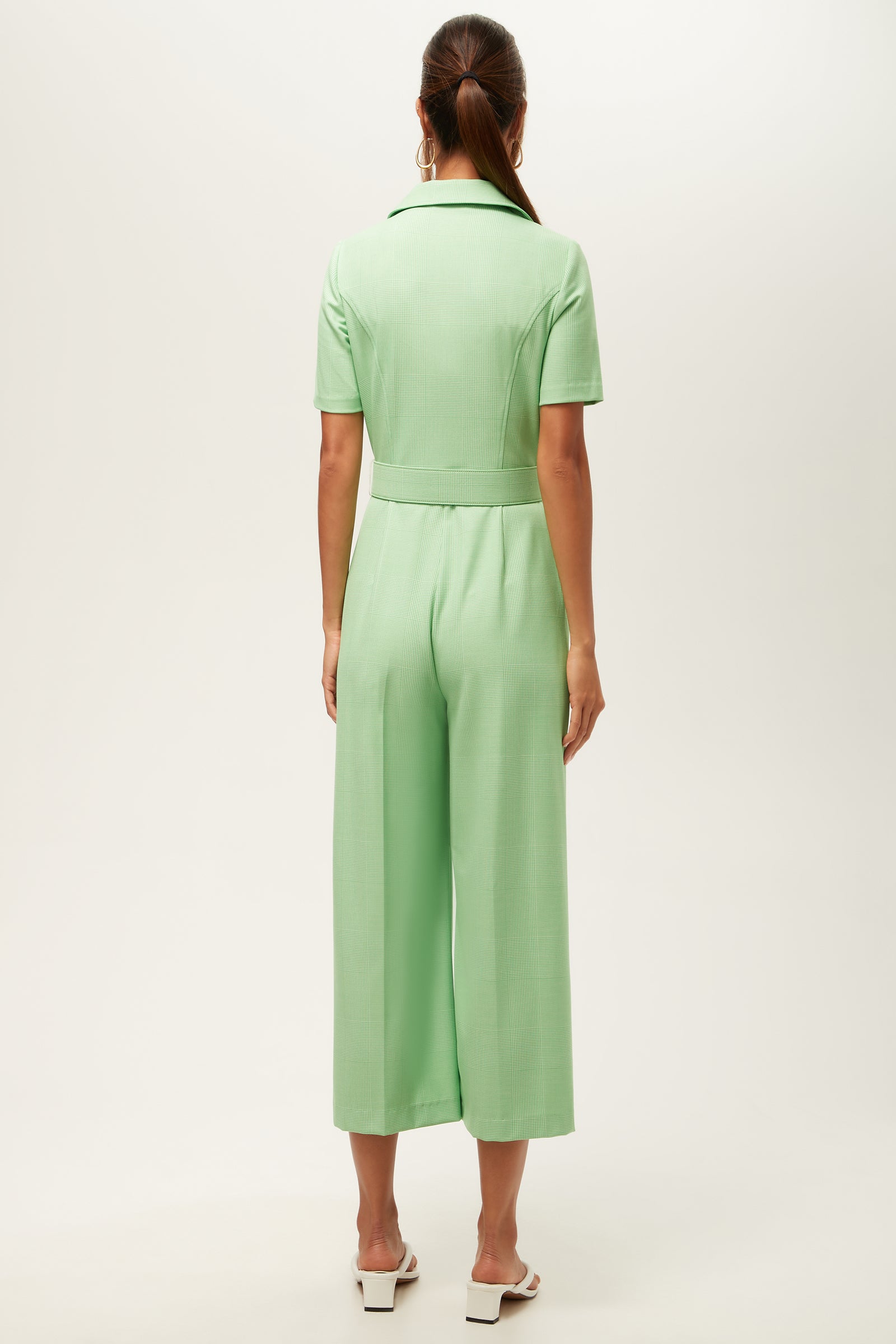 A woman with her hair tied back stands against a plain background, wearing the Jannise 3 Jumpsuit in Game Time Glen Plaid—a light green, short-sleeve, wide-leg jumpsuit with a collar—paired with white heeled sandals.
