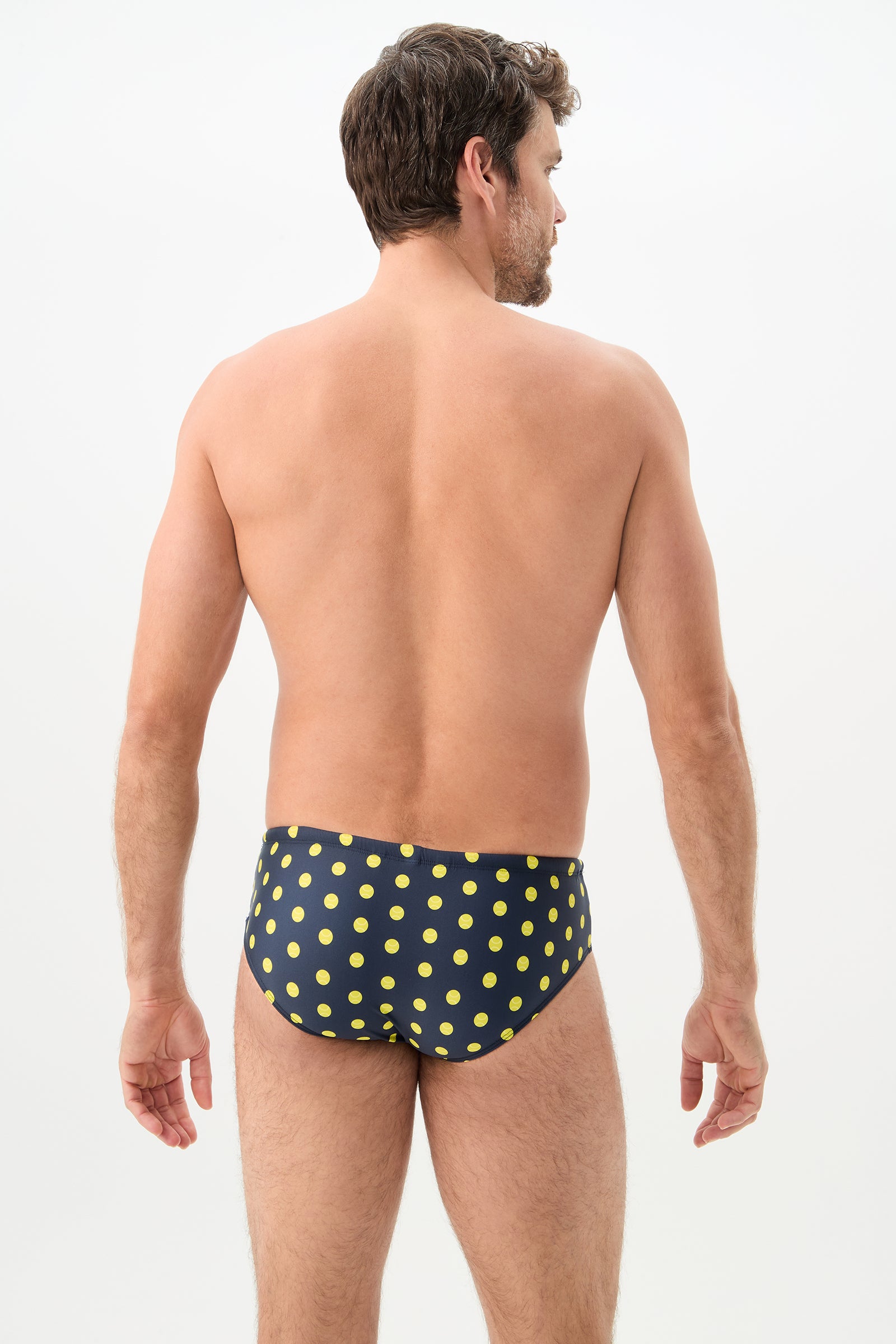A man faces away from the camera, wearing the Bondi Swim Brief - Tennis Polka Dot, a navy blue swim brief with yellow polka dots, set against a plain light background.