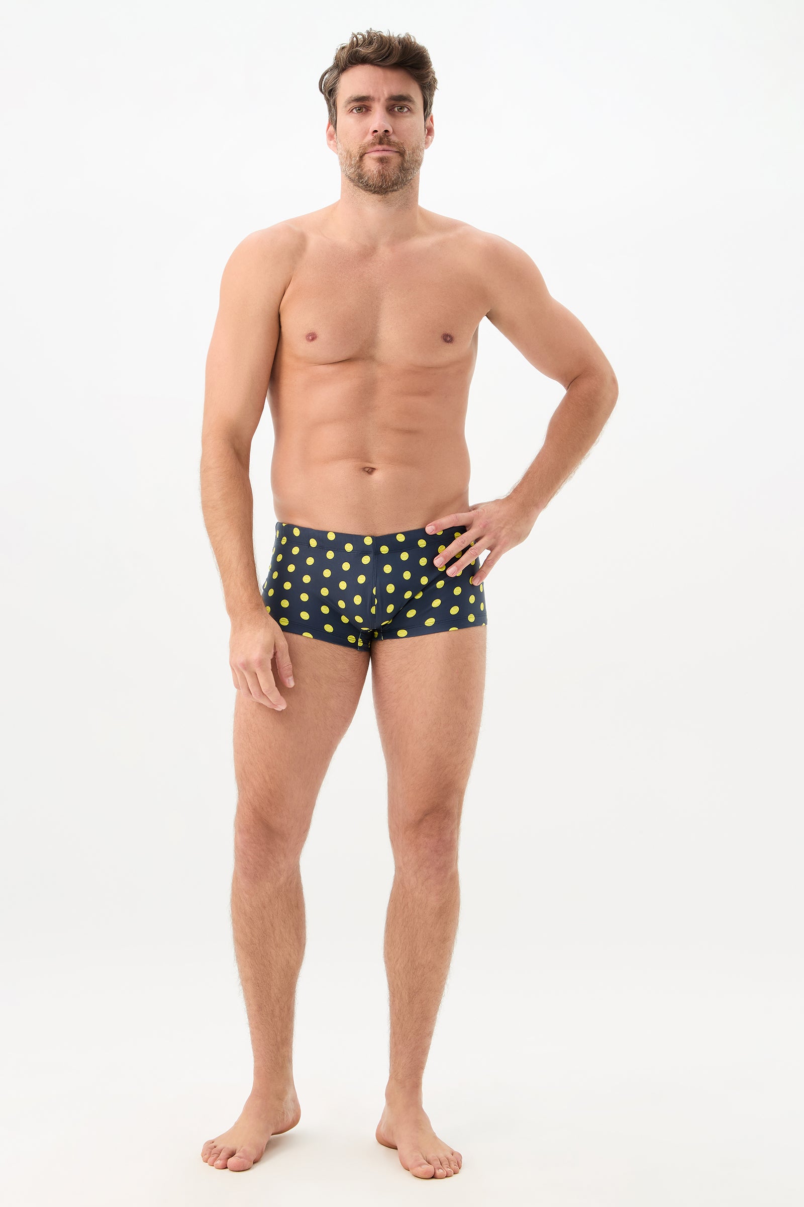 A man stands barefoot against a plain white background, wearing the Sorrento Swim Trunk in Tennis Polka Dot. He rests one hand on his hip and looks forward with a neutral expression.