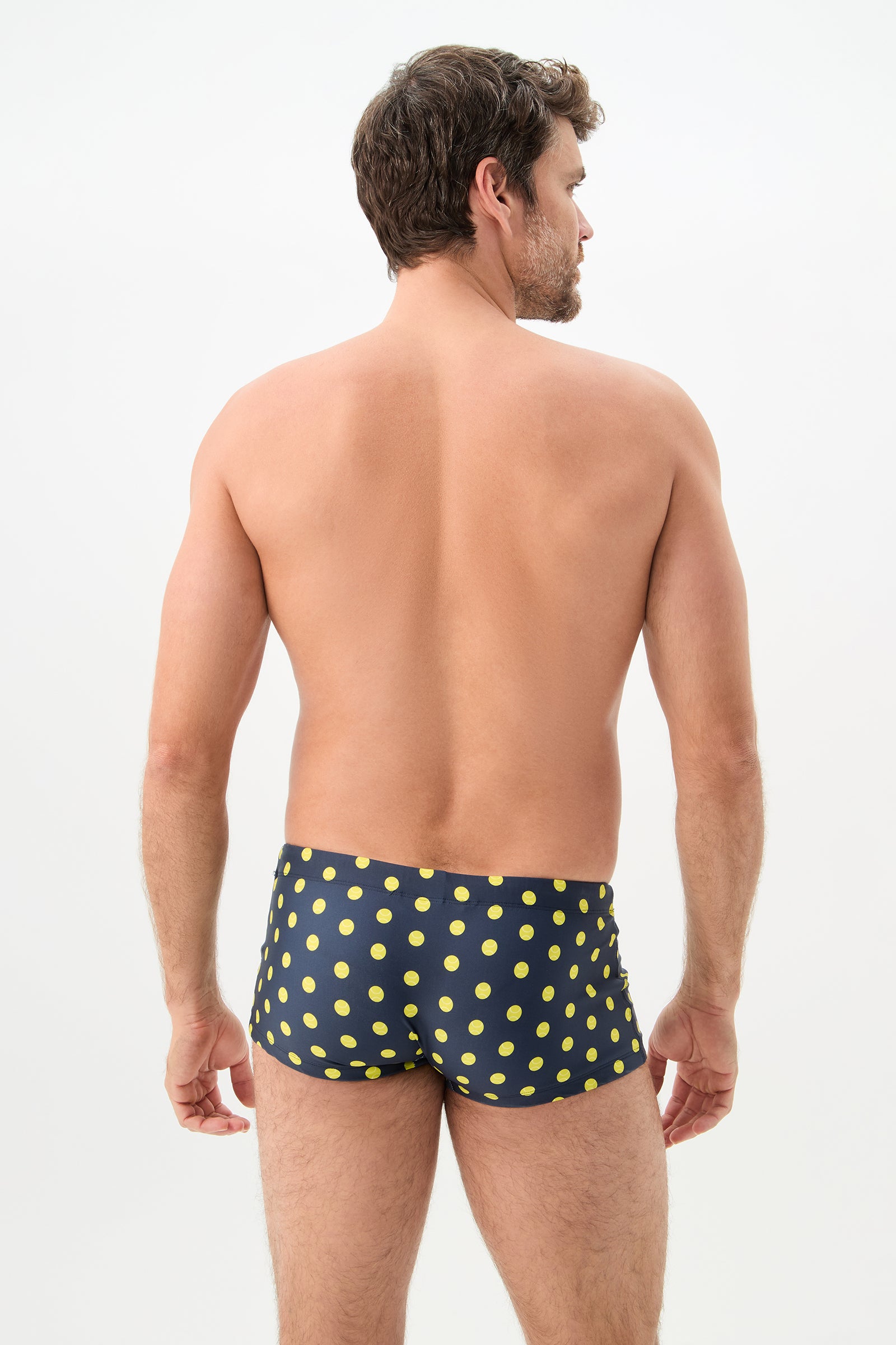 A man faces away from the camera, revealing his bare back and wearing the Sorrento Swim Trunk - Tennis Polka Dot. The swim trunks are navy blue with a yellow polka dot design, set against a plain light background.