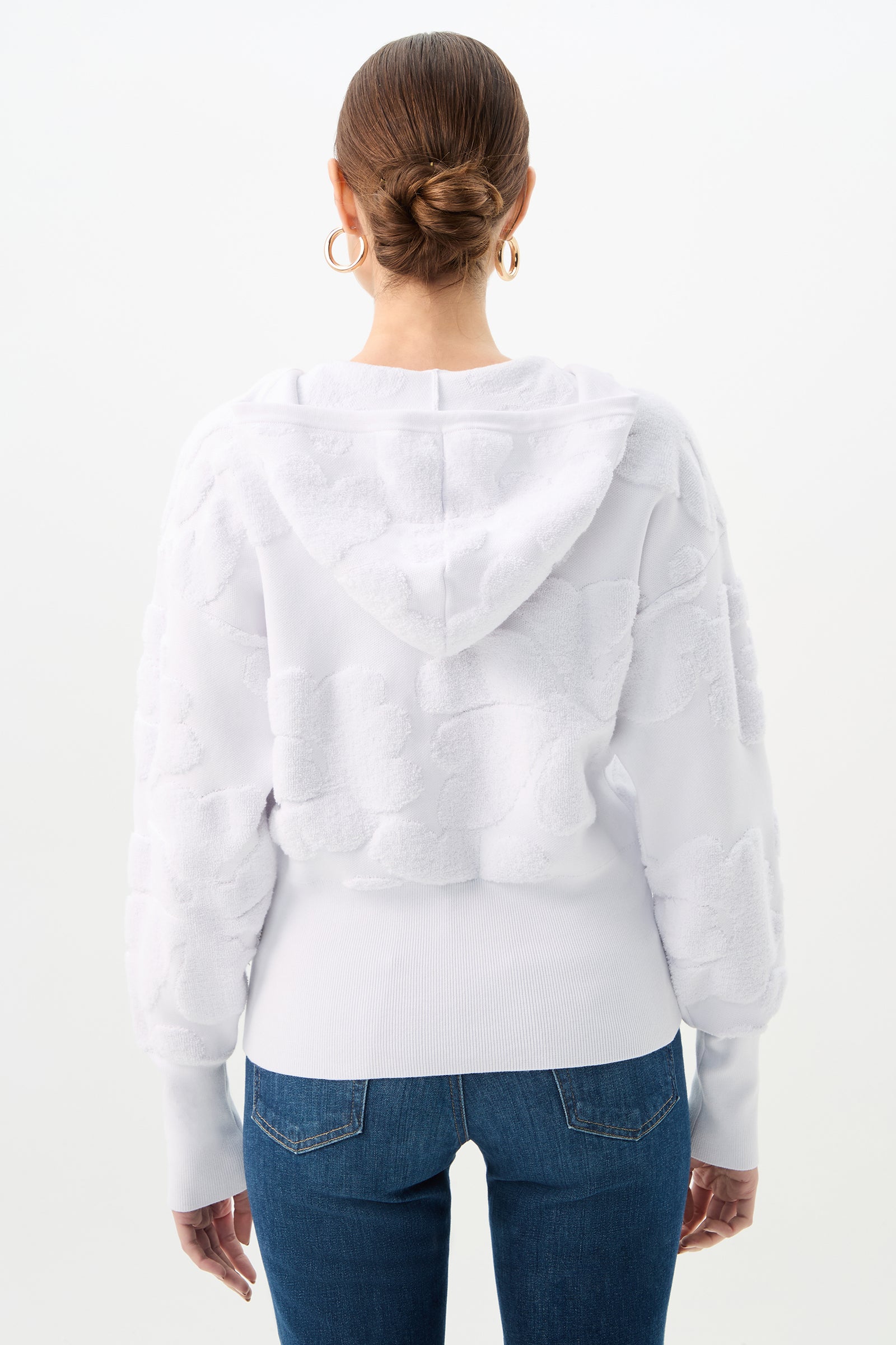 Woman with brown hair in a neat bun, wearing gold hoop earrings, blue jeans, and the C.Z. Zip-Up Hoodie - Linsk Terry in white, shown from the back against a plain white background.