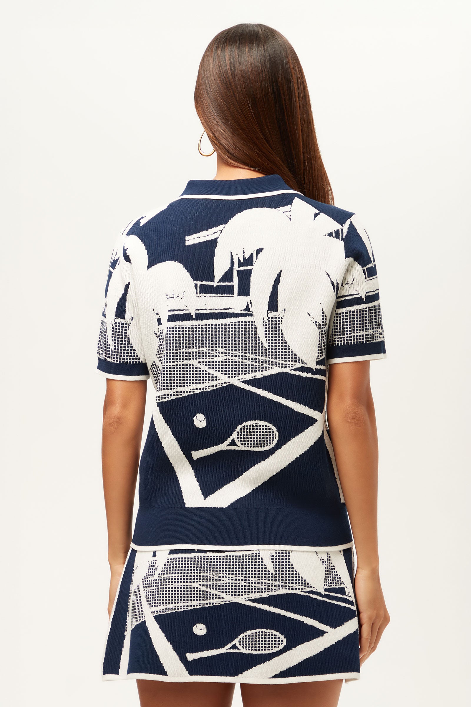 A woman with long brown hair, seen from behind, wears the La Jolla 2 Short-Sleeve Graphic Knit Polo featuring a navy and white tennis court, racquet, ball, and palm tree design. The background is plain white.