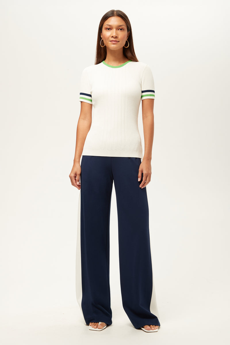 A woman faces forward against a plain white background, wearing the Record Wide-Leg Pant - Cozy Ponte in navy with a short-sleeve white ribbed top, green and blue striped sleeves, white heeled sandals, and hoop earrings.