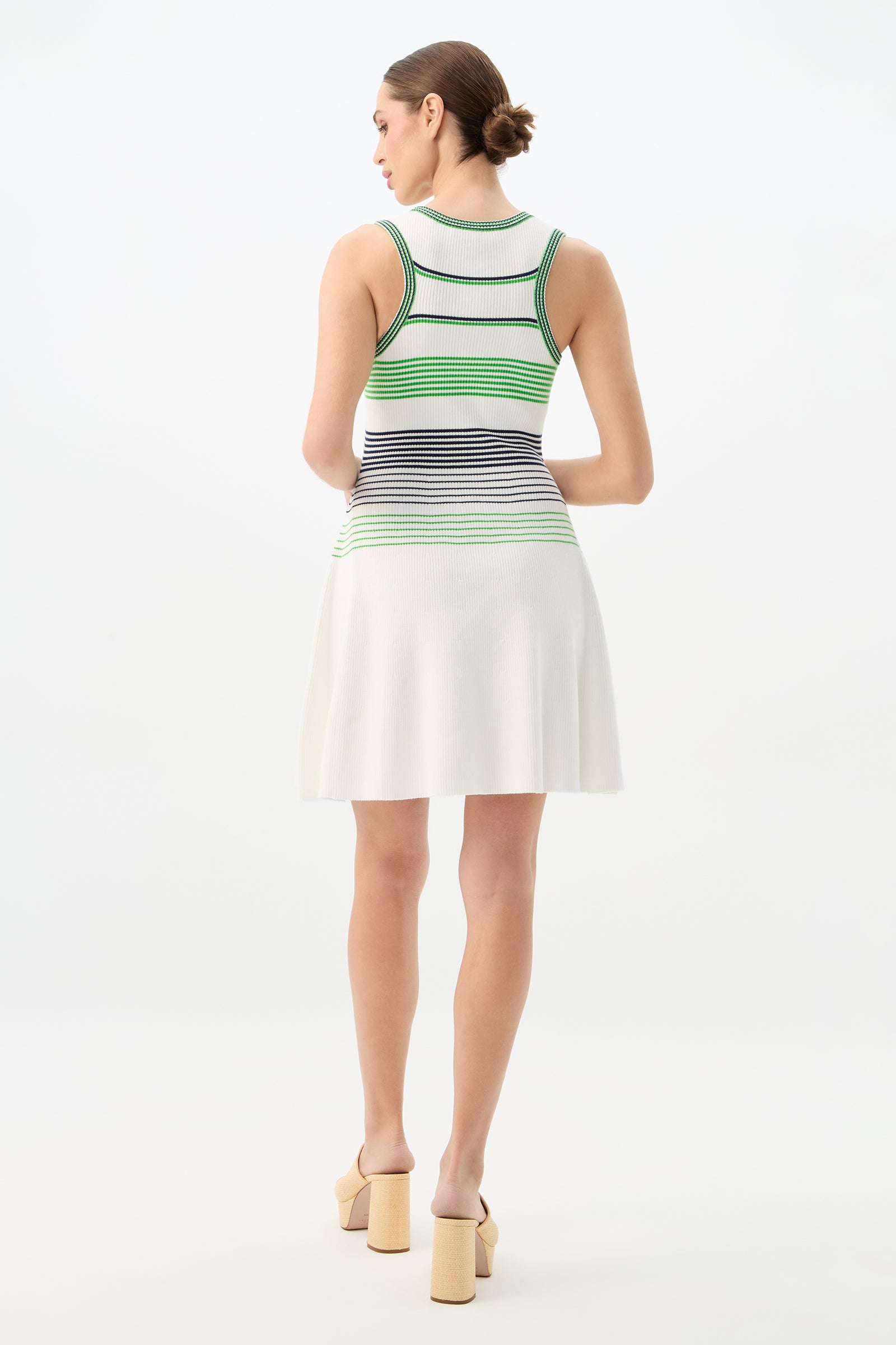 A woman faces away in the Veronika Sleeveless Rib Knit Sweater Dress, featuring green and black striped ribbing on top with a white skirt. Her hair is styled in a low bun, and she completes the look with beige platform heels.