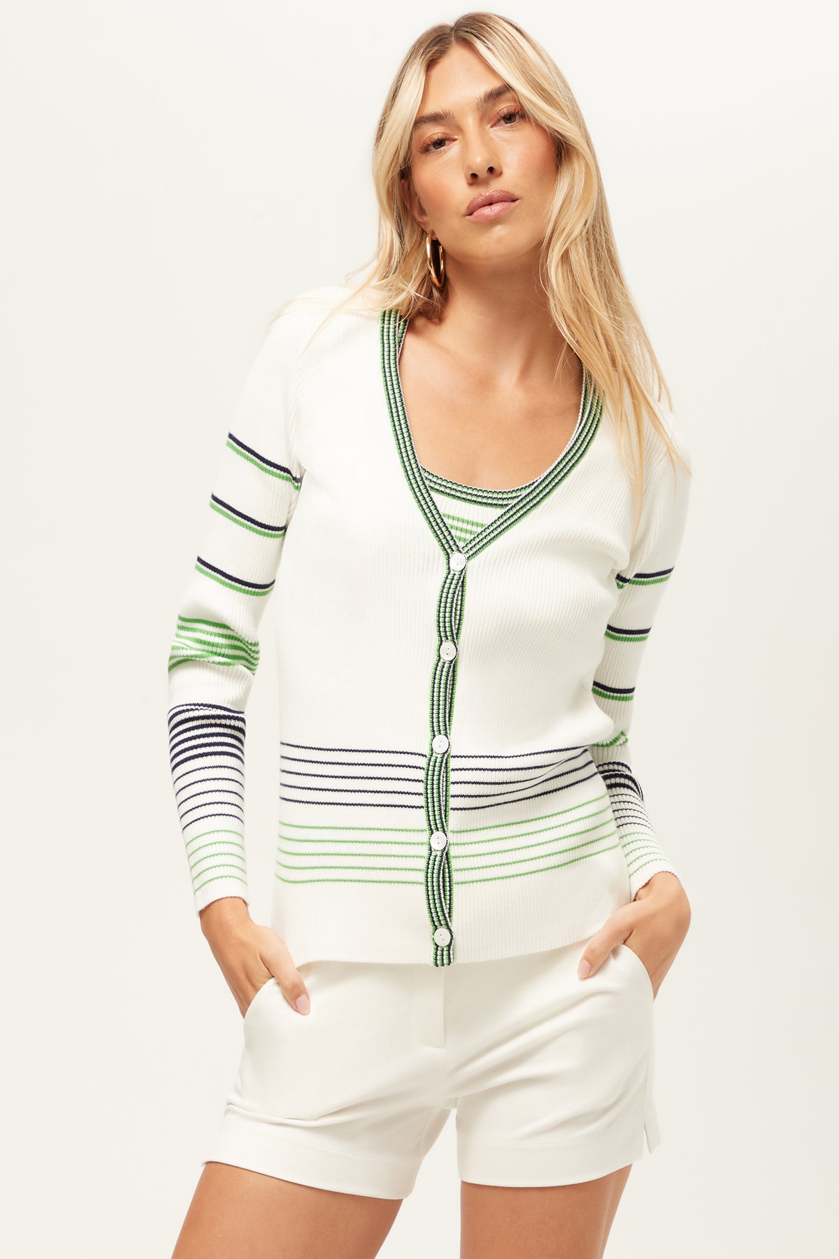 A woman with long blonde hair wears the Leylah V-Neck Striped Cardigan in Whitewash/Ink, paired with a matching striped top and white shorts, standing with her hands in her pockets against a plain background.