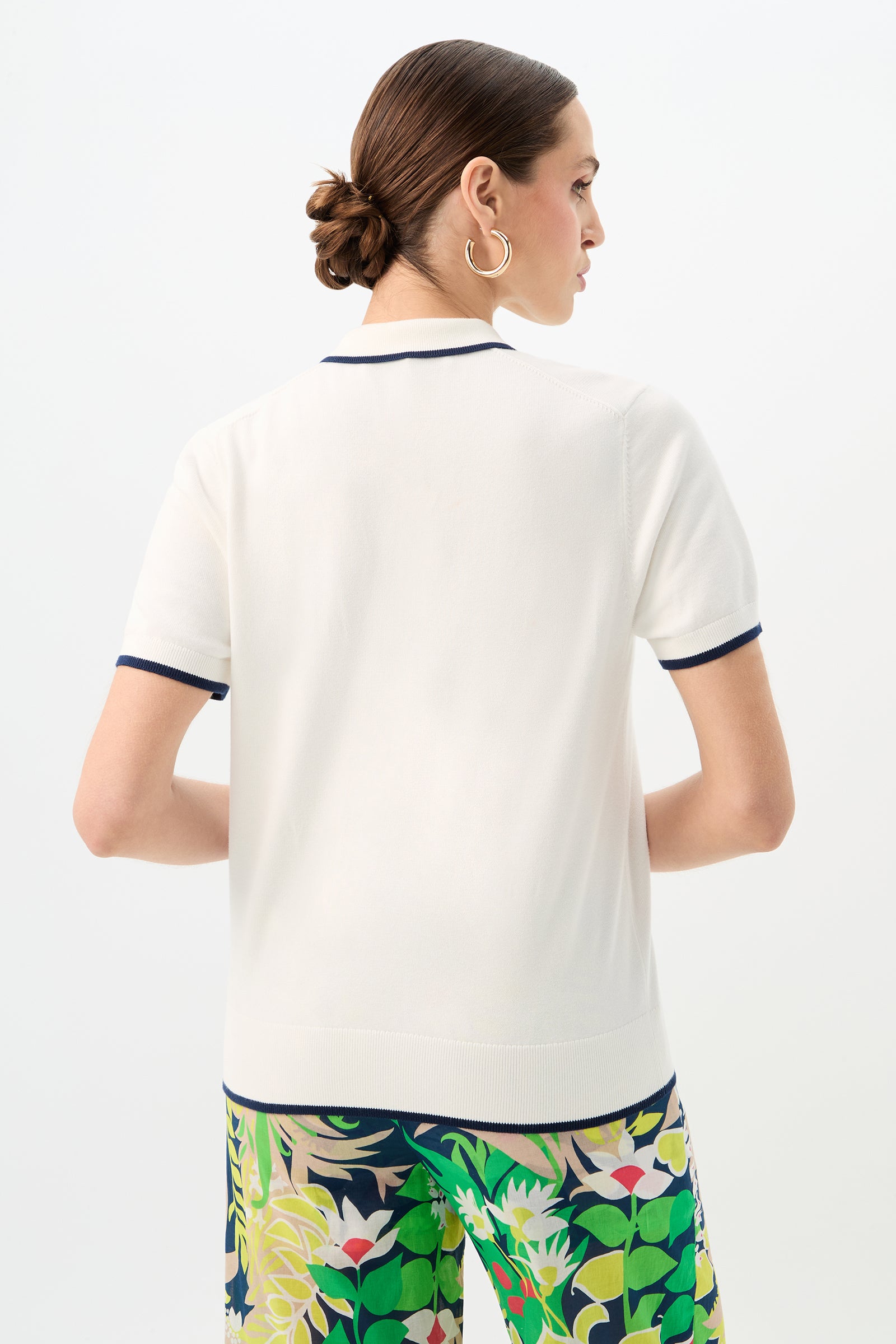 A woman with brown hair in a low bun, wearing the Serena Tennis Heart Short-Sleeve Polo, large hoop earrings, and colorful floral pants, faces away from the camera against a plain background.