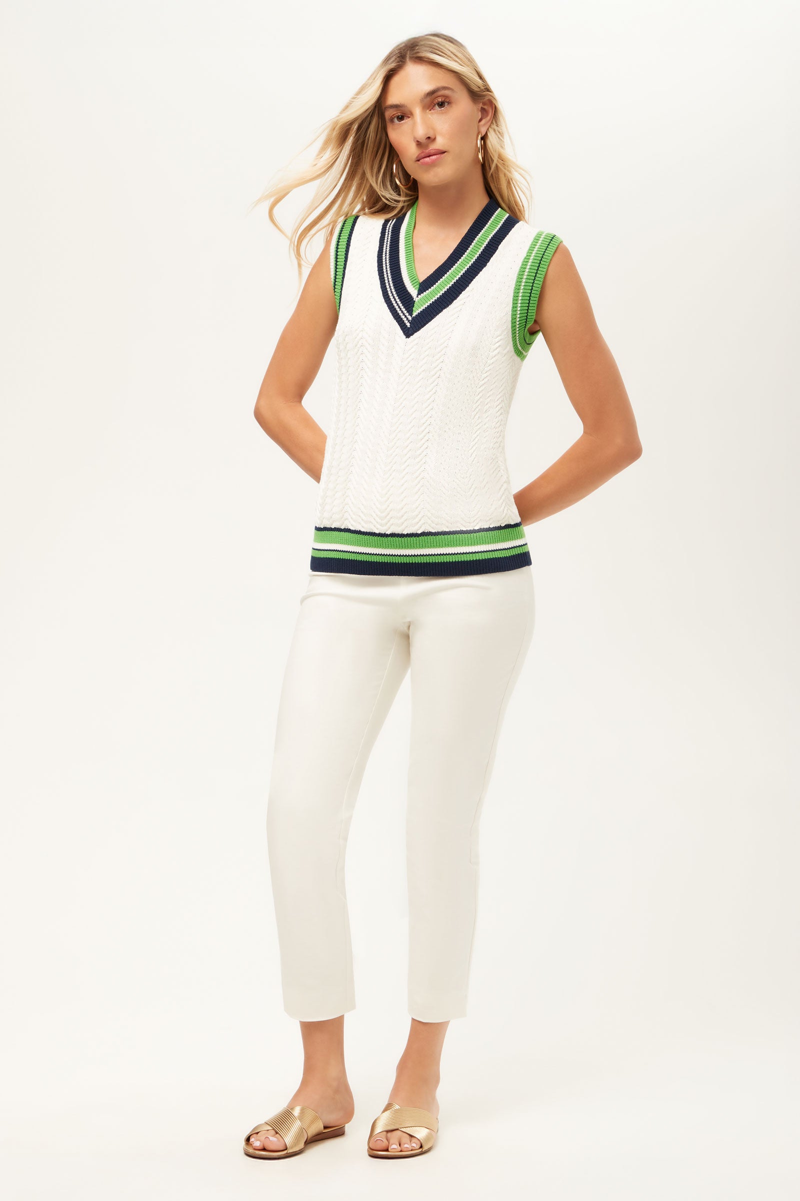 A woman wearing the Seapointe 2 Sleeveless Knit Vest, featuring white cable-knit with green and navy trim, stands against a white background. She pairs it with white slim pants and gold sandals, her long blonde hair down and expression neutral.