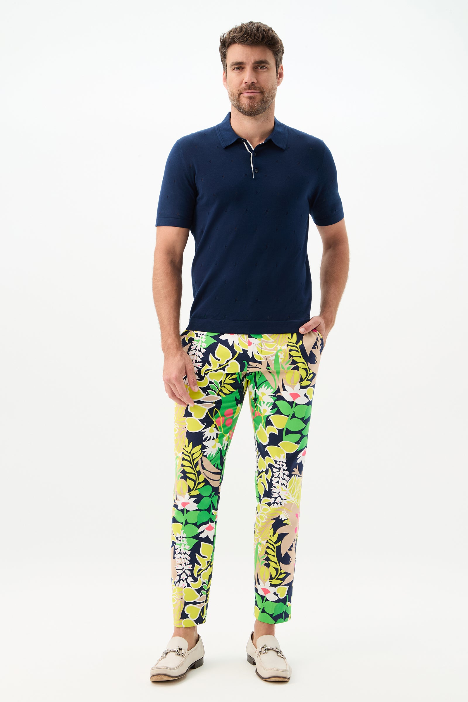 A man stands against a white background wearing the Clyde Slim Trouser - PS Eternal, a navy blue polo shirt, and beige loafers, with one hand in his pocket and a relaxed expression.