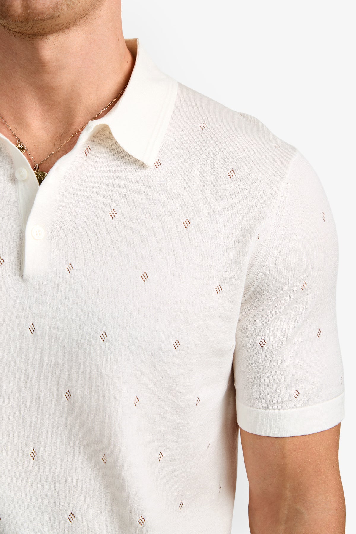 A person is shown from the upper torso up, wearing the Nicholas Short-Sleeve Polo—a white knit shirt featuring a collar and small diamond-shaped perforated patterns—against a plain background.