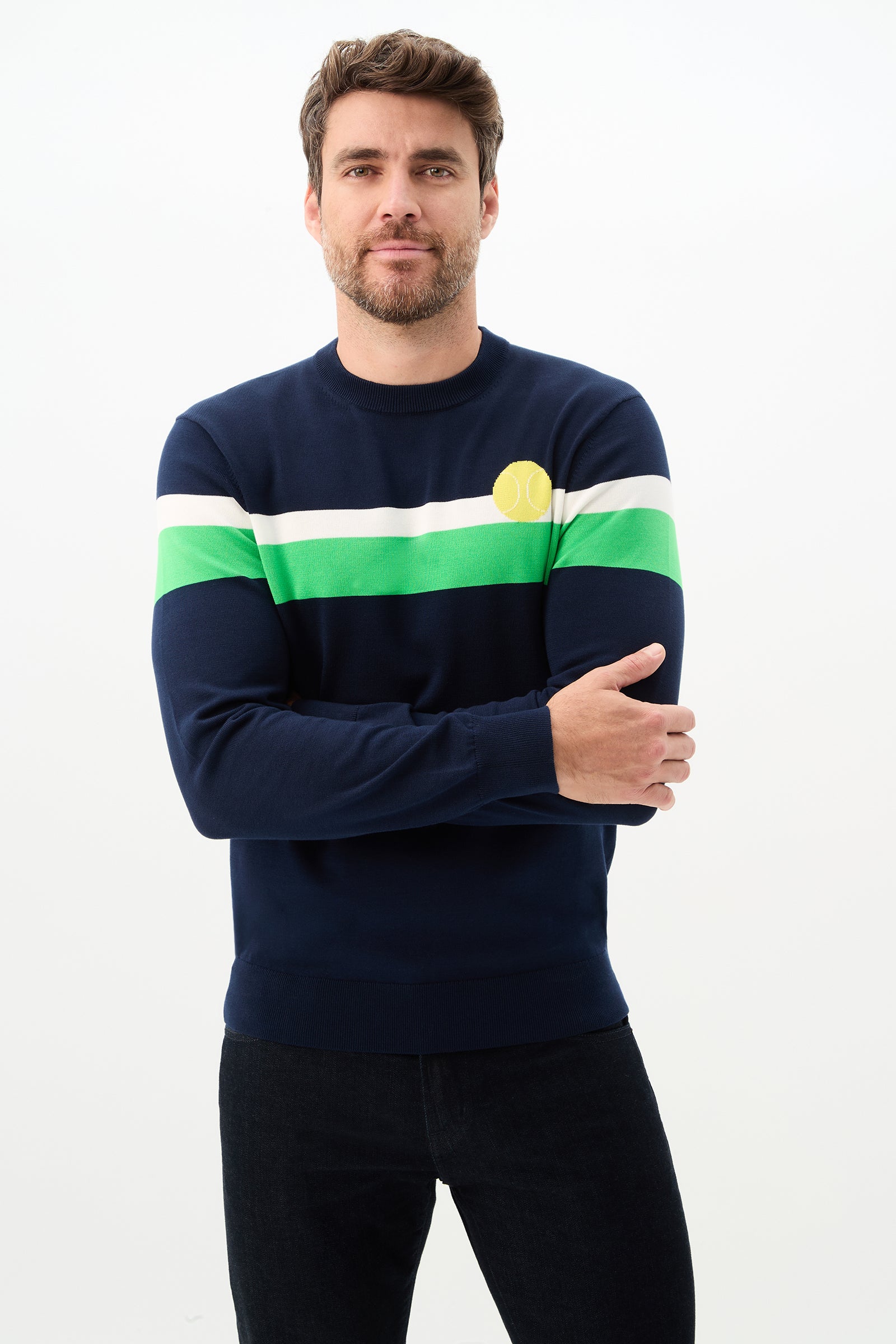 A man with short brown hair and a beard wears the Romero Crewneck Sweater in Ink, featuring white and green horizontal stripes and a yellow tennis ball emblem, standing against a plain white background.