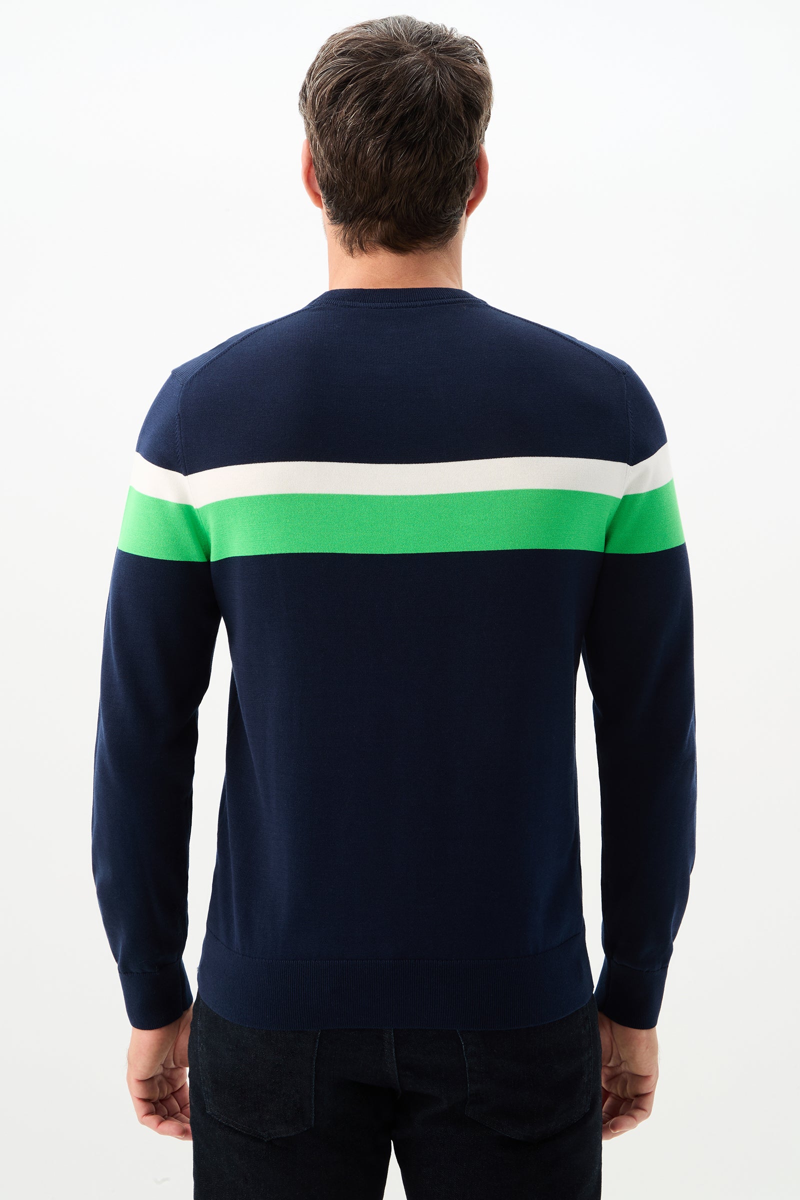 A man with short brown hair stands with his back to the camera, wearing dark pants and the Romero Crewneck Sweater—navy blue with white and green stripes across the upper back and sleeves.