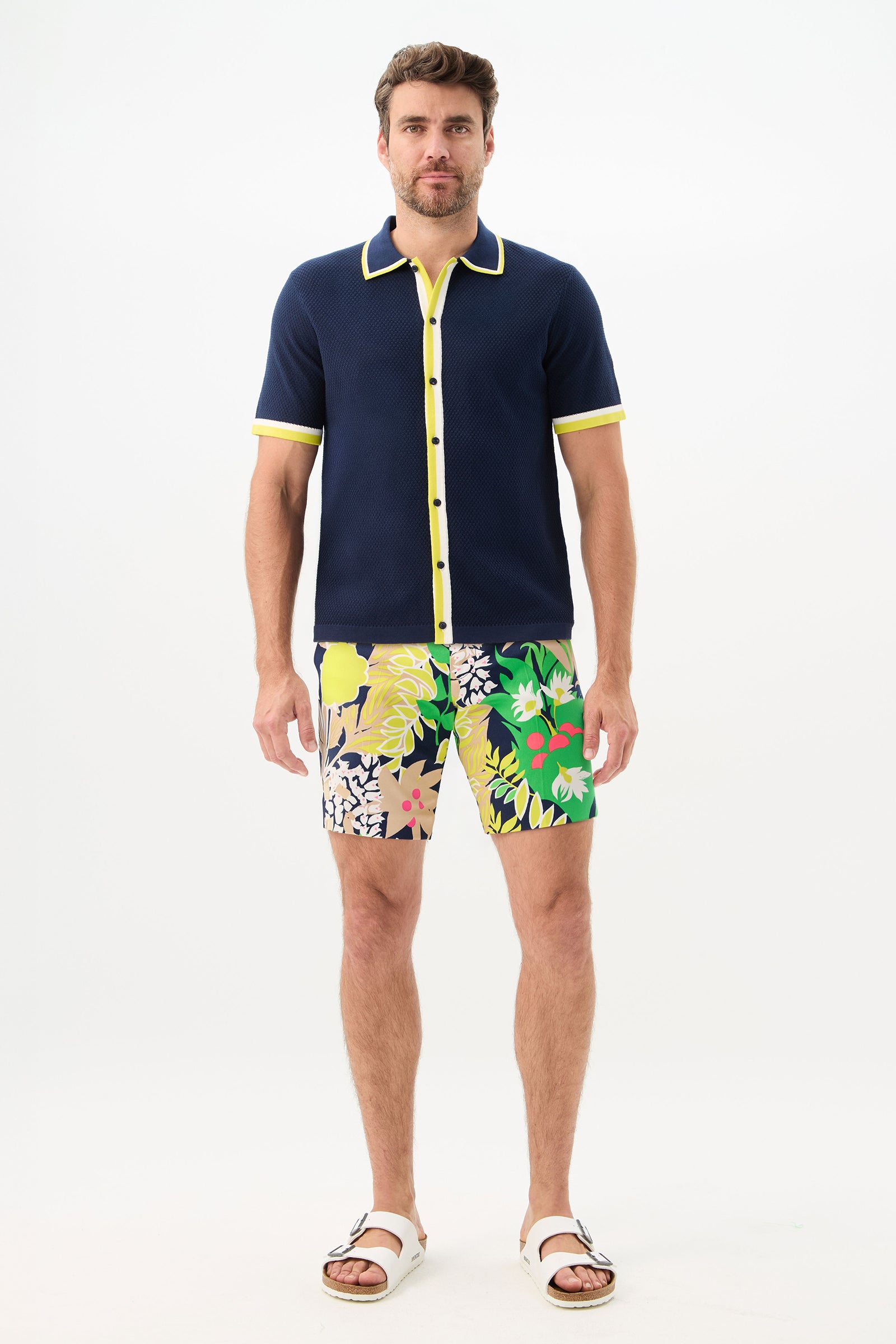 A man faces forward wearing the Lawrence Short - PS Eternal, a navy short-sleeve shirt with yellow trim, colorful floral shorts, and white sandals against a plain white background.