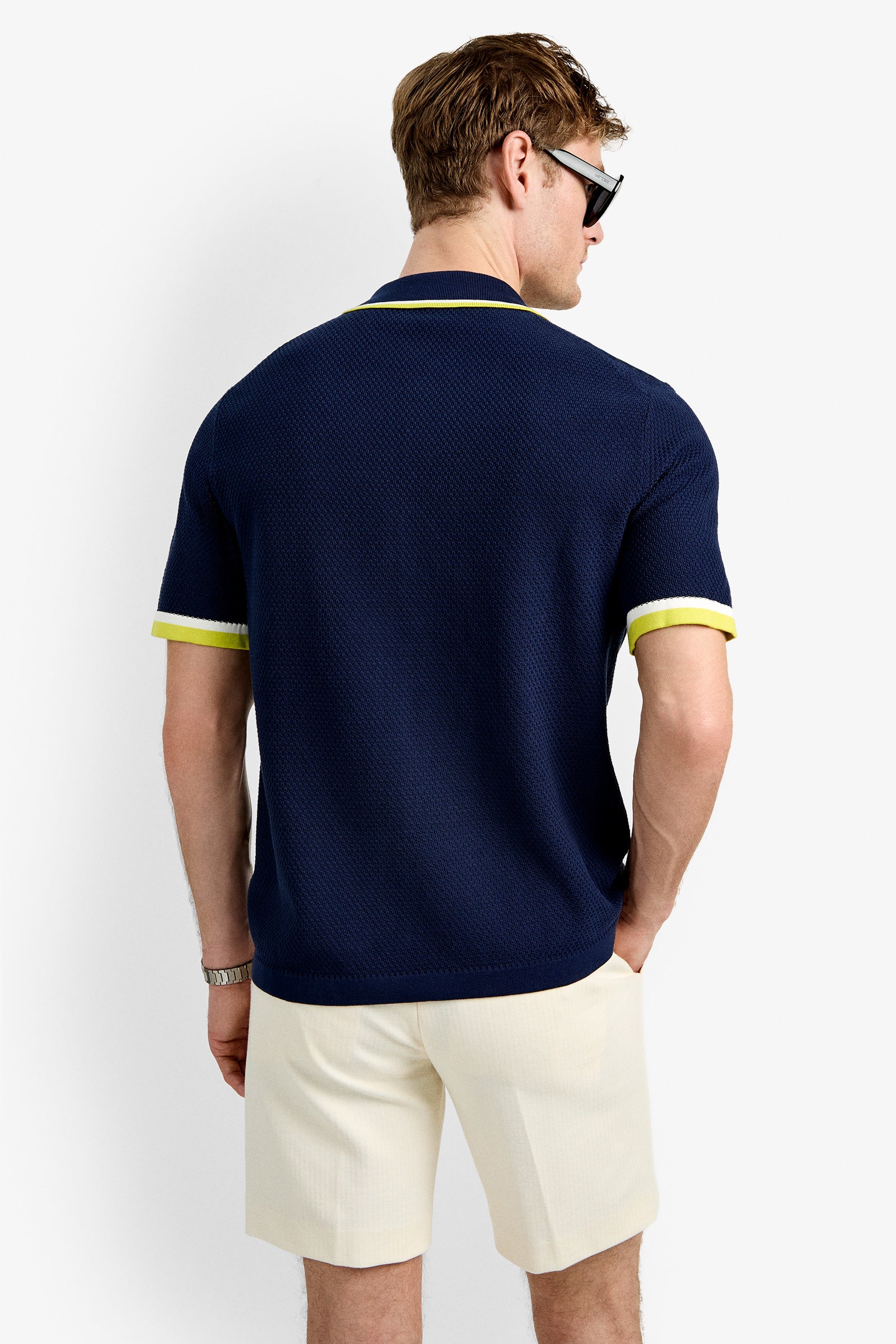 A man in sunglasses wears the Kylian Short-Sleeve Polo—navy blue with yellow trim—and cream shorts, standing with one hand in his pocket against a plain white background.
