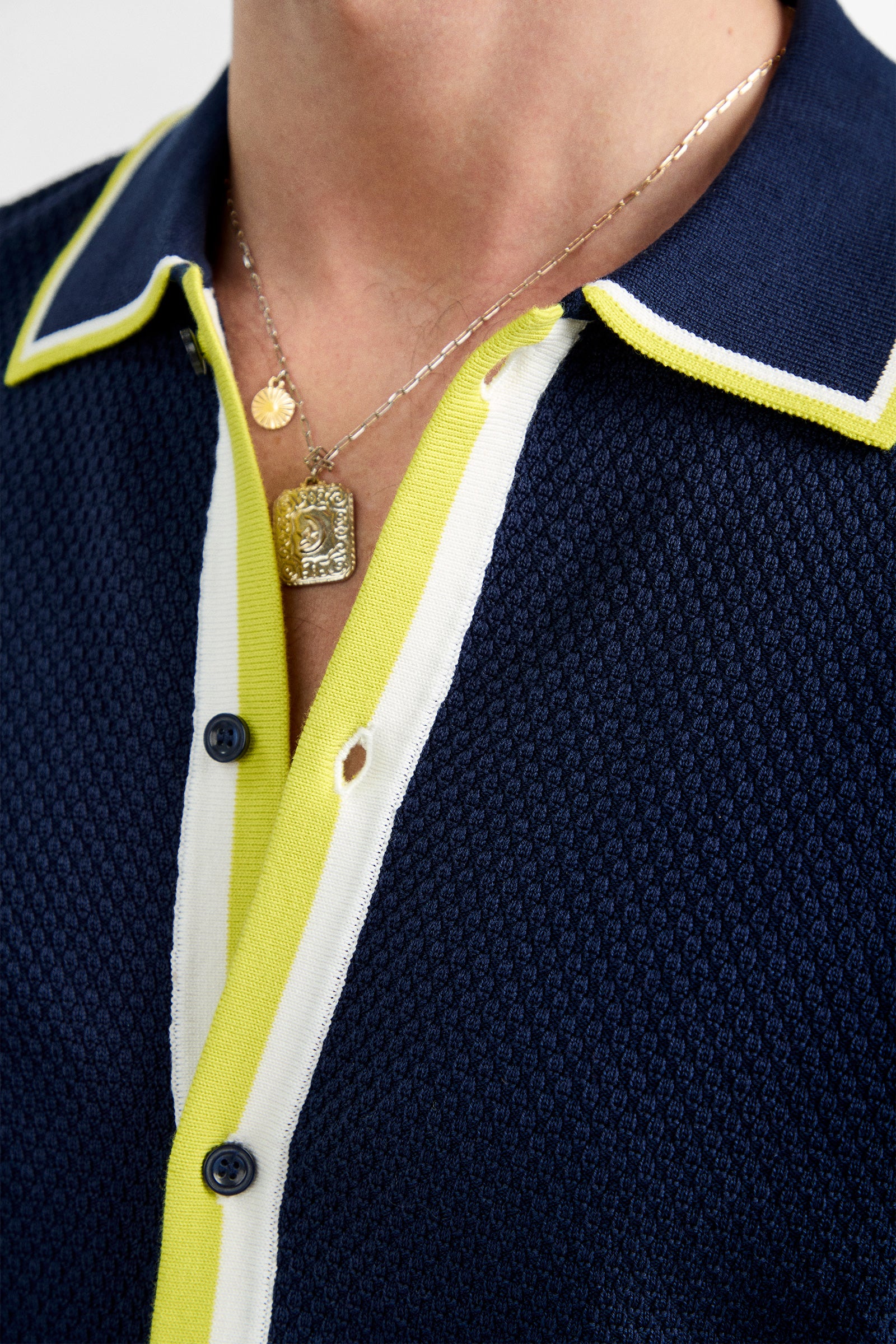 Close-up of a person wearing the Kylian Short-Sleeve Polo in navy blue with yellow and white trim, blue buttons, and layered gold necklaces featuring round and rectangular pendants.