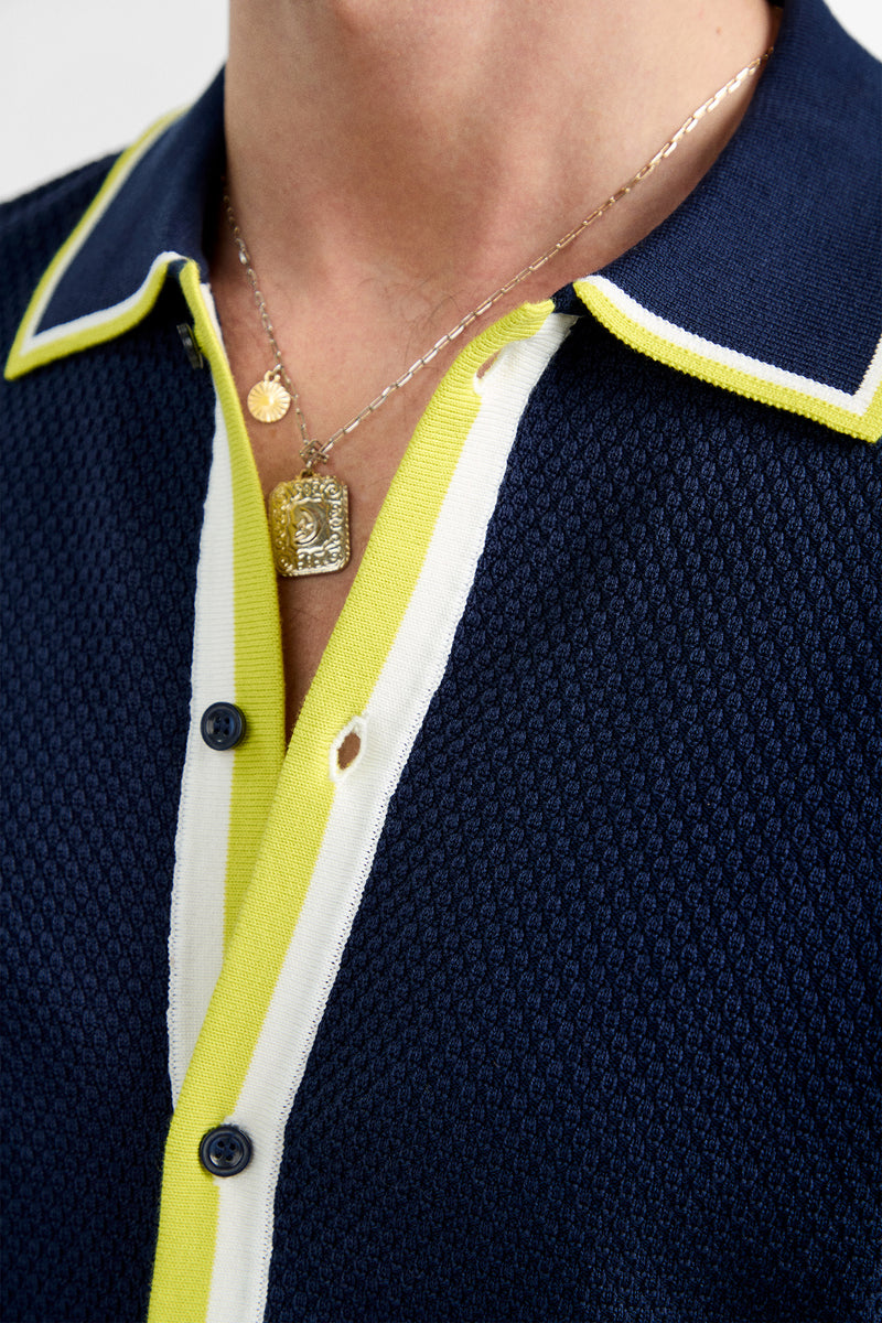 Close-up of a person wearing the Kylian Short-Sleeve Polo in navy blue with yellow and white trim, blue buttons, and layered gold necklaces featuring round and rectangular pendants.