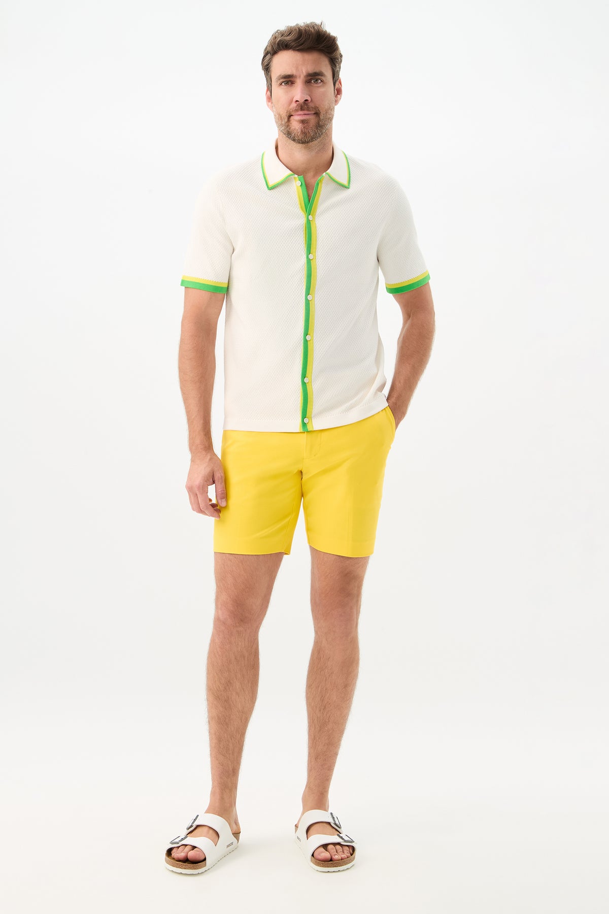 A man stands against a white background wearing the Cyrus Short - Sunset Stretch in yellow, paired with a white short-sleeve shirt featuring green trim and white sandals. One hand is in his pocket as he looks straight ahead.