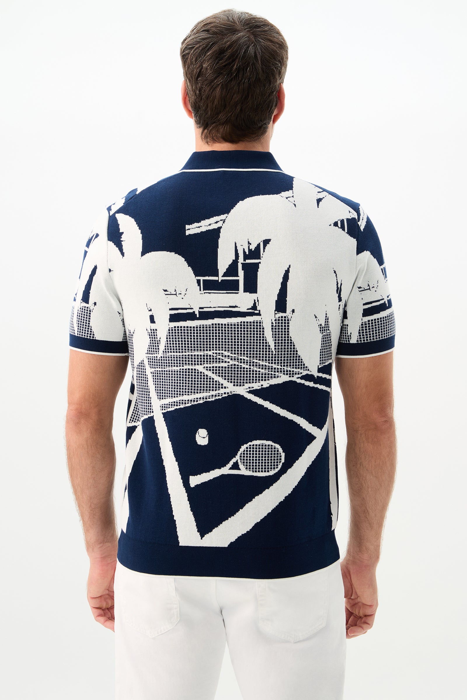 A man wears the Grayson Short-Sleeve Polo - Graphic Knit, featuring a navy blue color with a graphic of a white tennis court, palm trees, and tennis equipment on the back, standing against a plain white background.