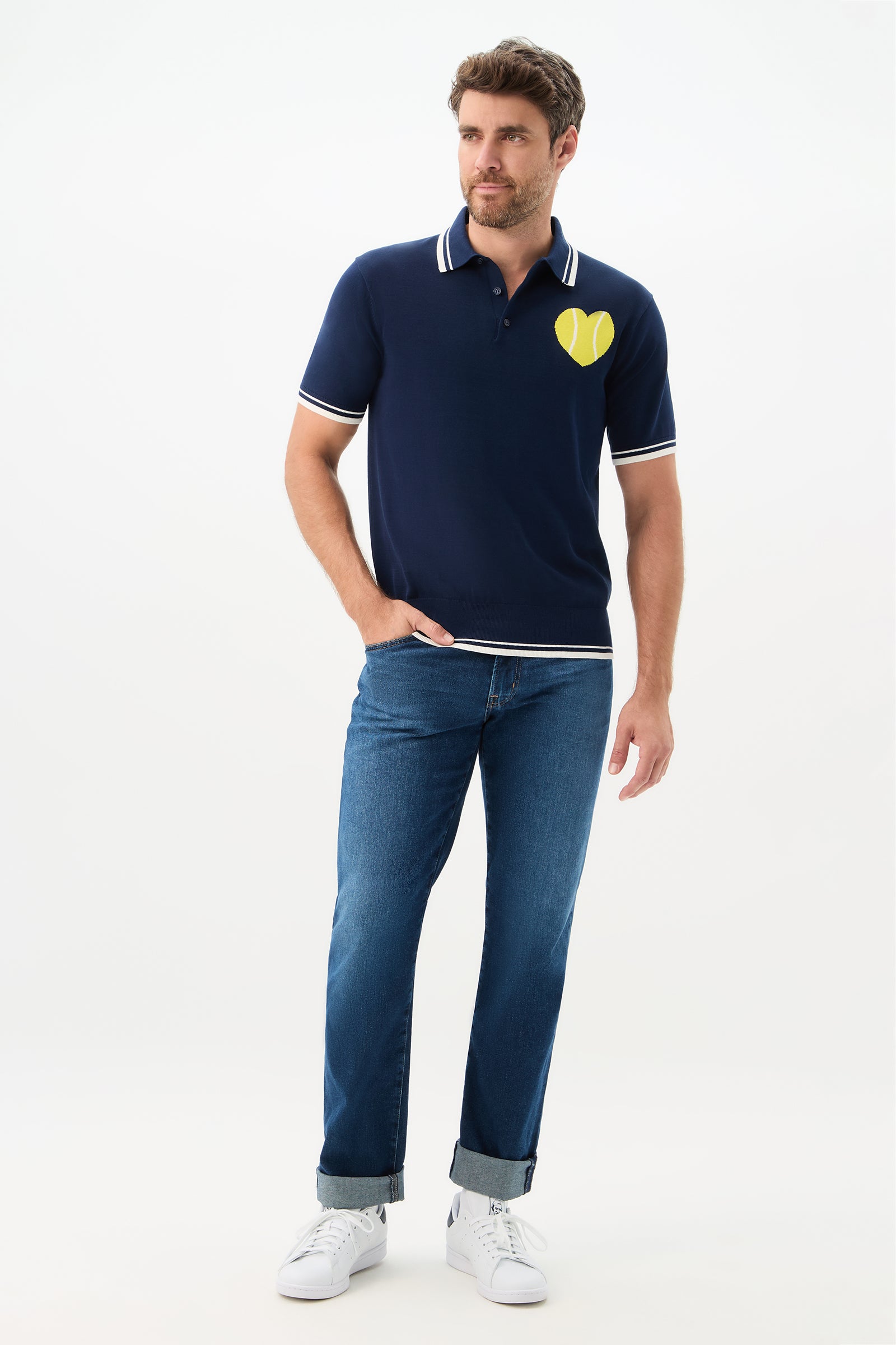 A man wears the Tennis Heart Short-Sleeve Polo—navy blue with a yellow tennis ball heart on the chest—paired with blue jeans and white sneakers, standing against a plain white background.