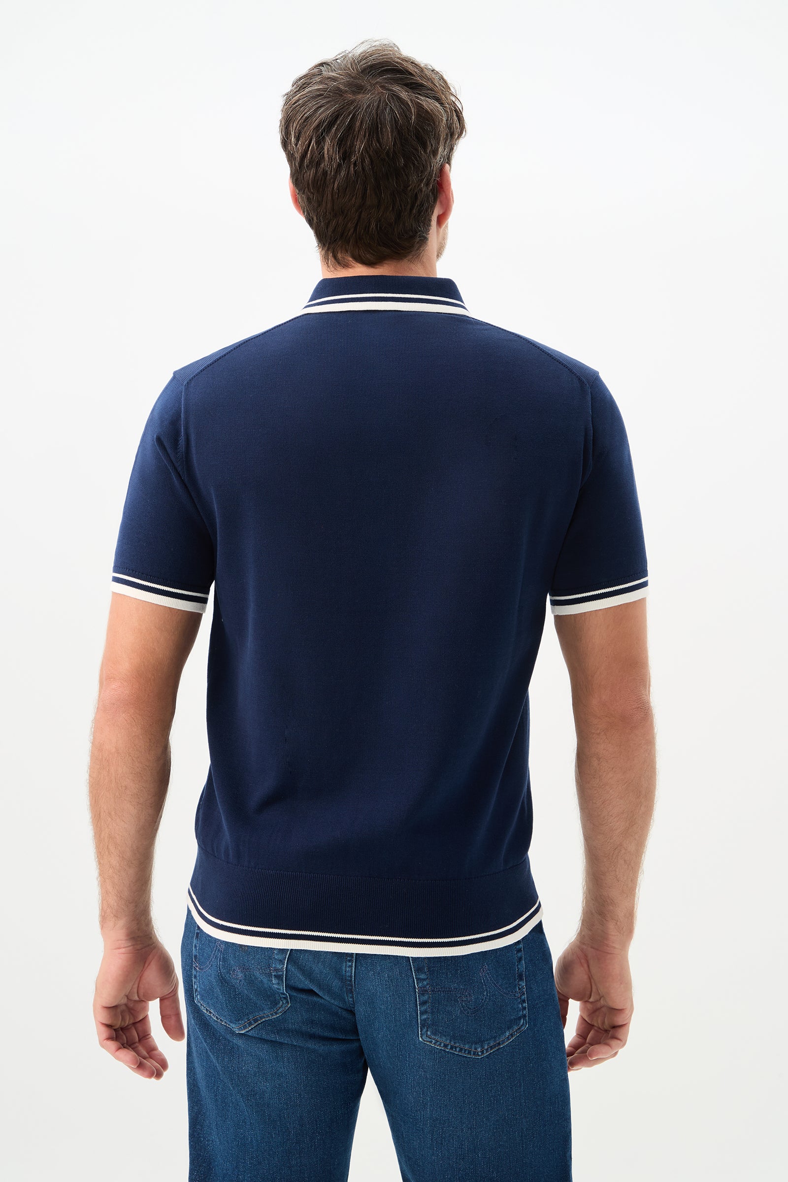 A man with short brown hair stands with his back to the camera, wearing the Tennis Heart Short-Sleeve Polo in navy blue with white trim and blue jeans against a plain light background.