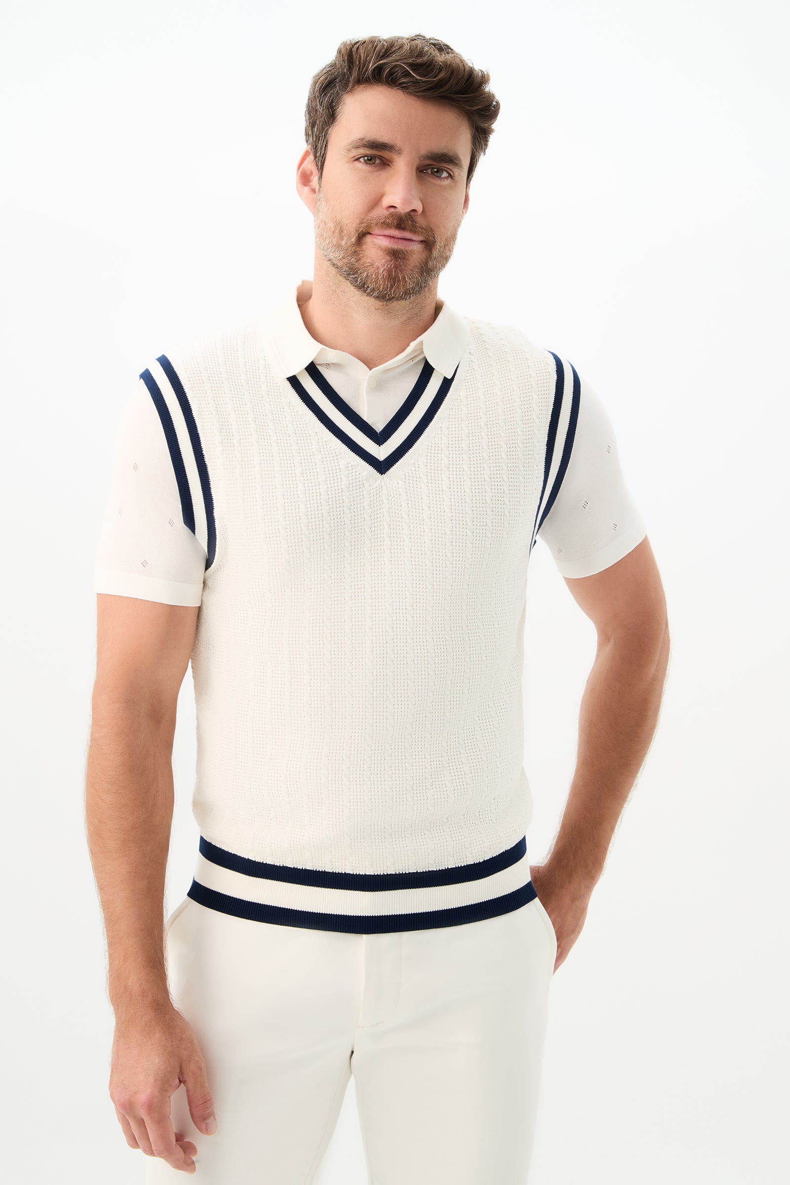 A man with short brown hair and a beard is wearing the Matthew V-Neck Knit Vest in Indigo/Optic White over a white collared shirt and white pants, standing against a plain light background.