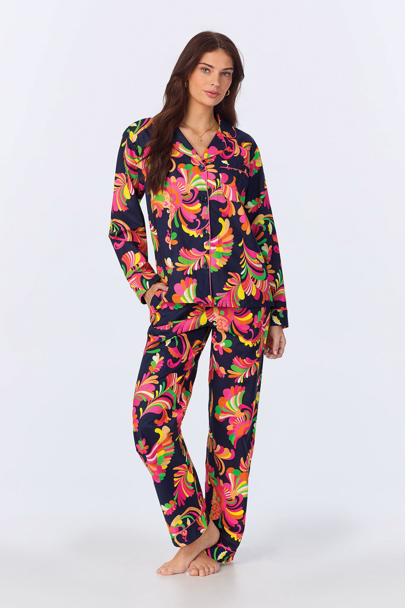 A woman with long brown hair stands barefoot, looking relaxed with one hand in her pocket, wearing the TT LS Pant PJ Set - Theodora Poplin in Multi, featuring vibrant florals on a dark background against a plain backdrop.