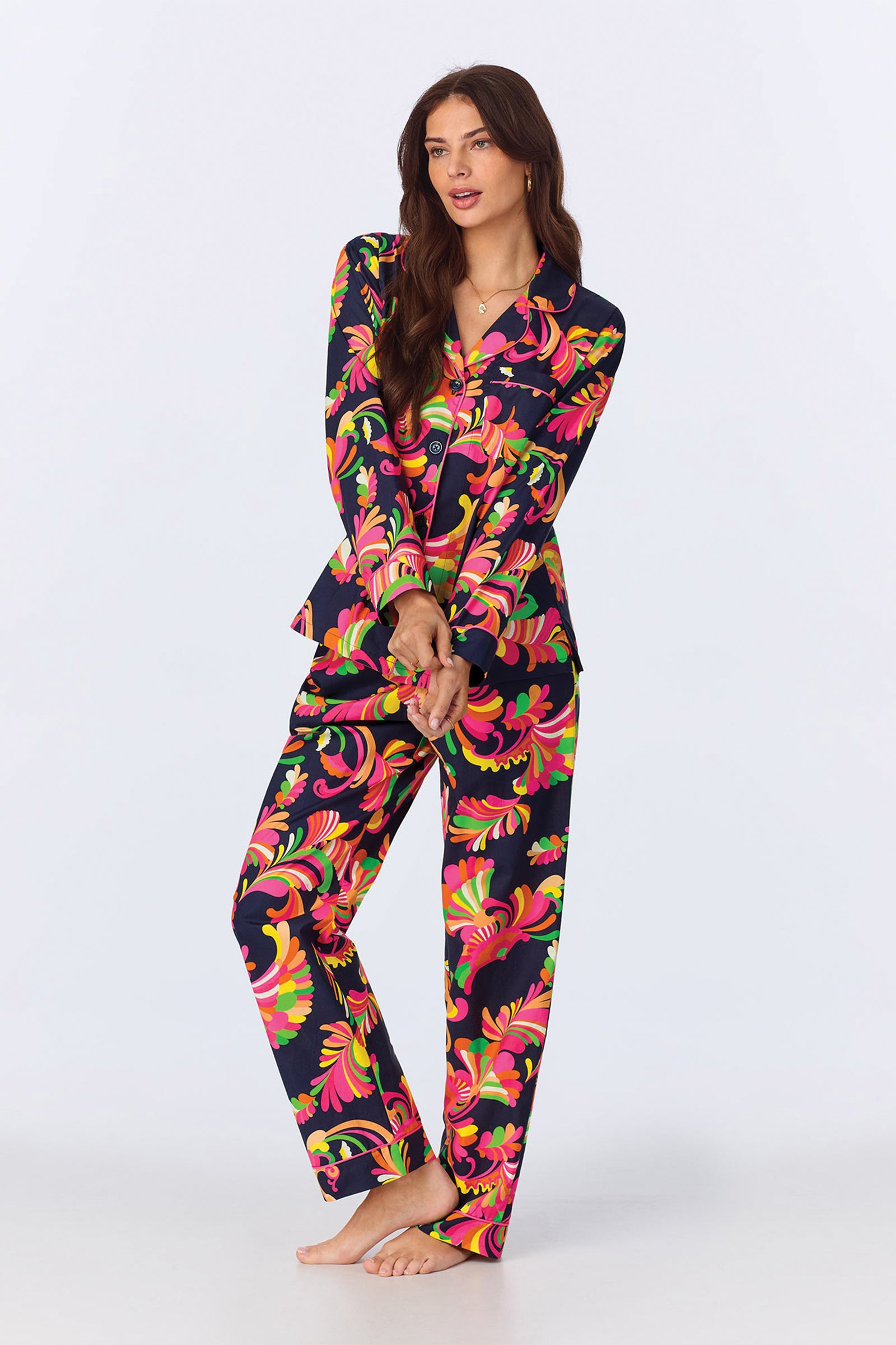 A woman with long brown hair wears the TT LS Pant PJ Set -Theodora Poplin, featuring a vibrant multicolored floral pattern on a dark background. She stands barefoot and poses against a plain light backdrop.