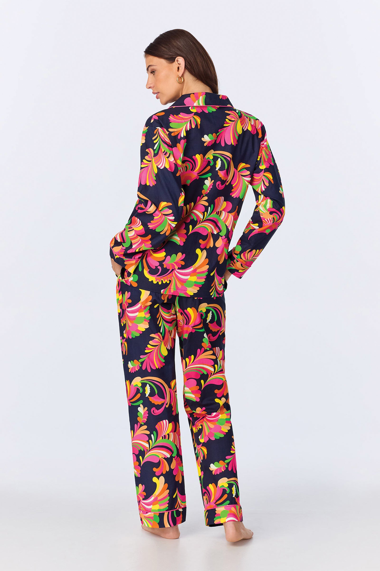 A person stands barefoot, facing away, wearing the TT LS Pant PJ Set - Theodora Poplin featuring matching long-sleeve pajamas and pants with a bold, swirling floral pattern on a dark background.