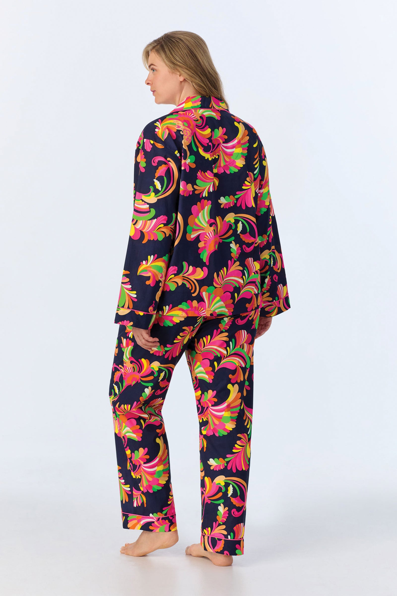 Wearing the TT LS Pant PJ Set - Theodora Poplin, a person stands barefoot and facing away in matching pajamas with a bold pink, orange, and green floral pattern on dark fabric. The background is plain white.