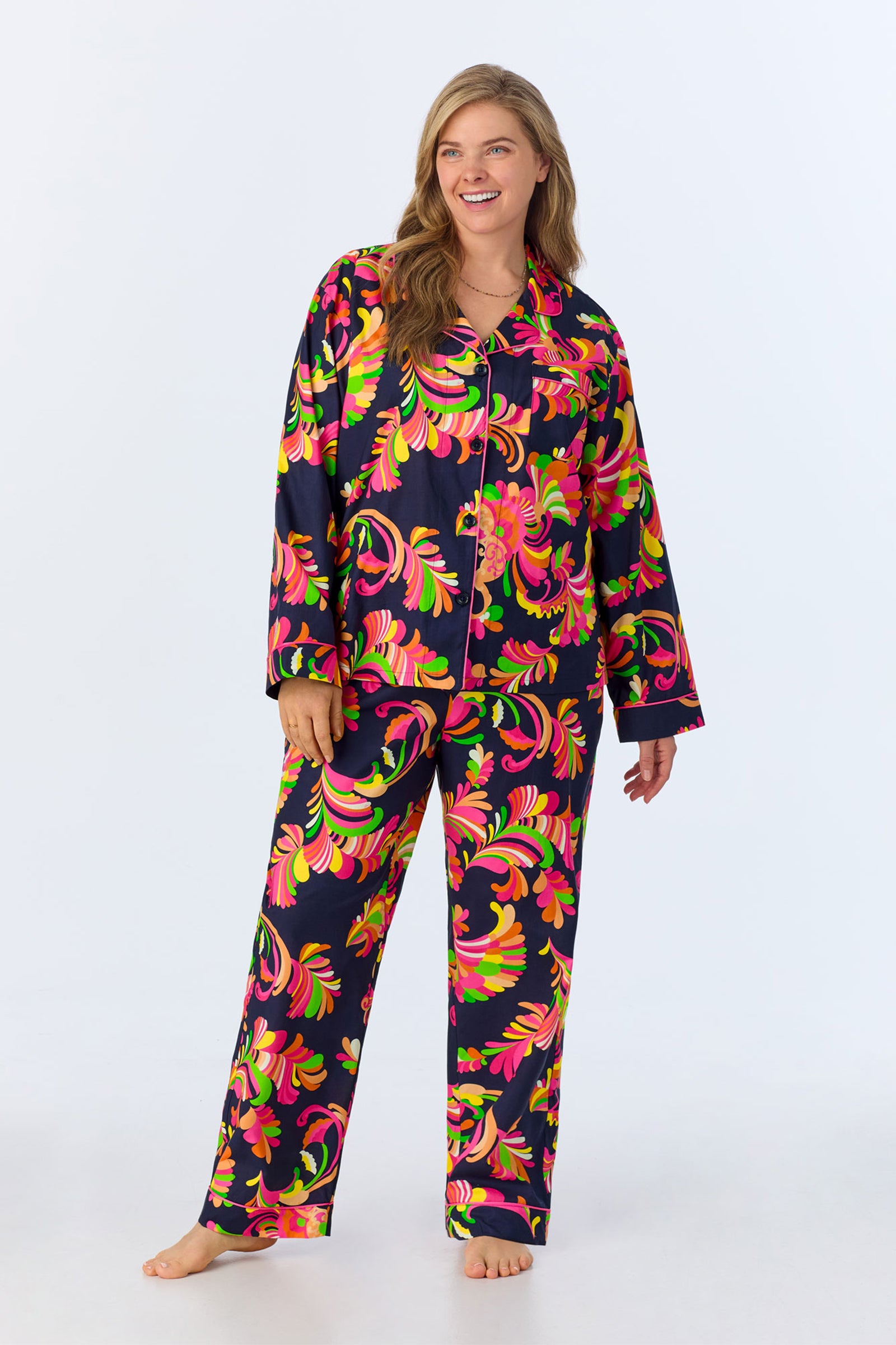 A woman smiles while wearing the TT LS Pant PJ Set - Theodora Poplin, featuring a bold swirl pattern in pink, green, and orange on a dark background. She stands barefoot against a plain, light backdrop.