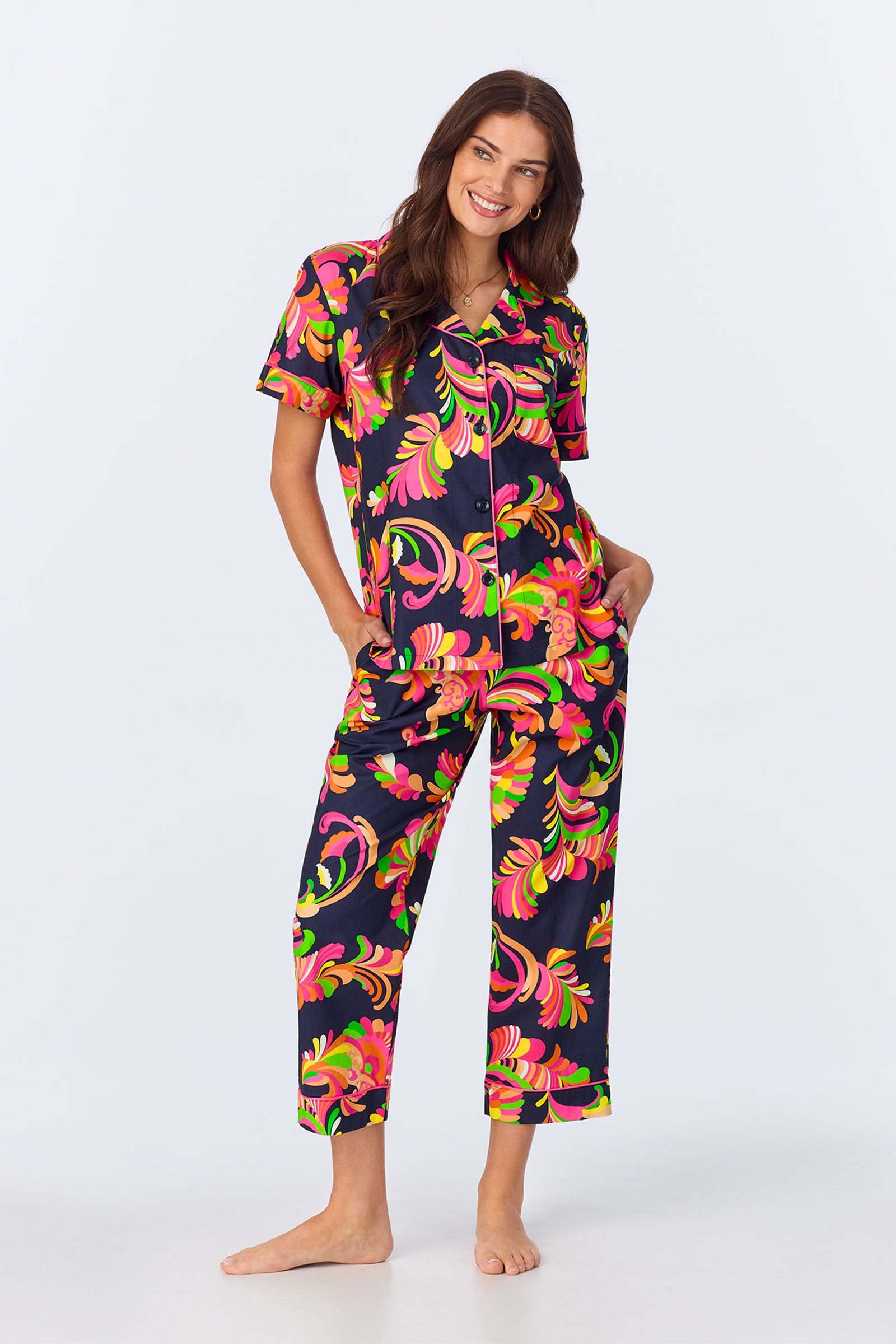 A woman smiles while wearing the TT SS Crop PJ Set - Theodora Poplin in Multi, featuring a matching short-sleeve button-up top and pants with an abstract floral print, standing barefoot with hands in pockets against a white background.