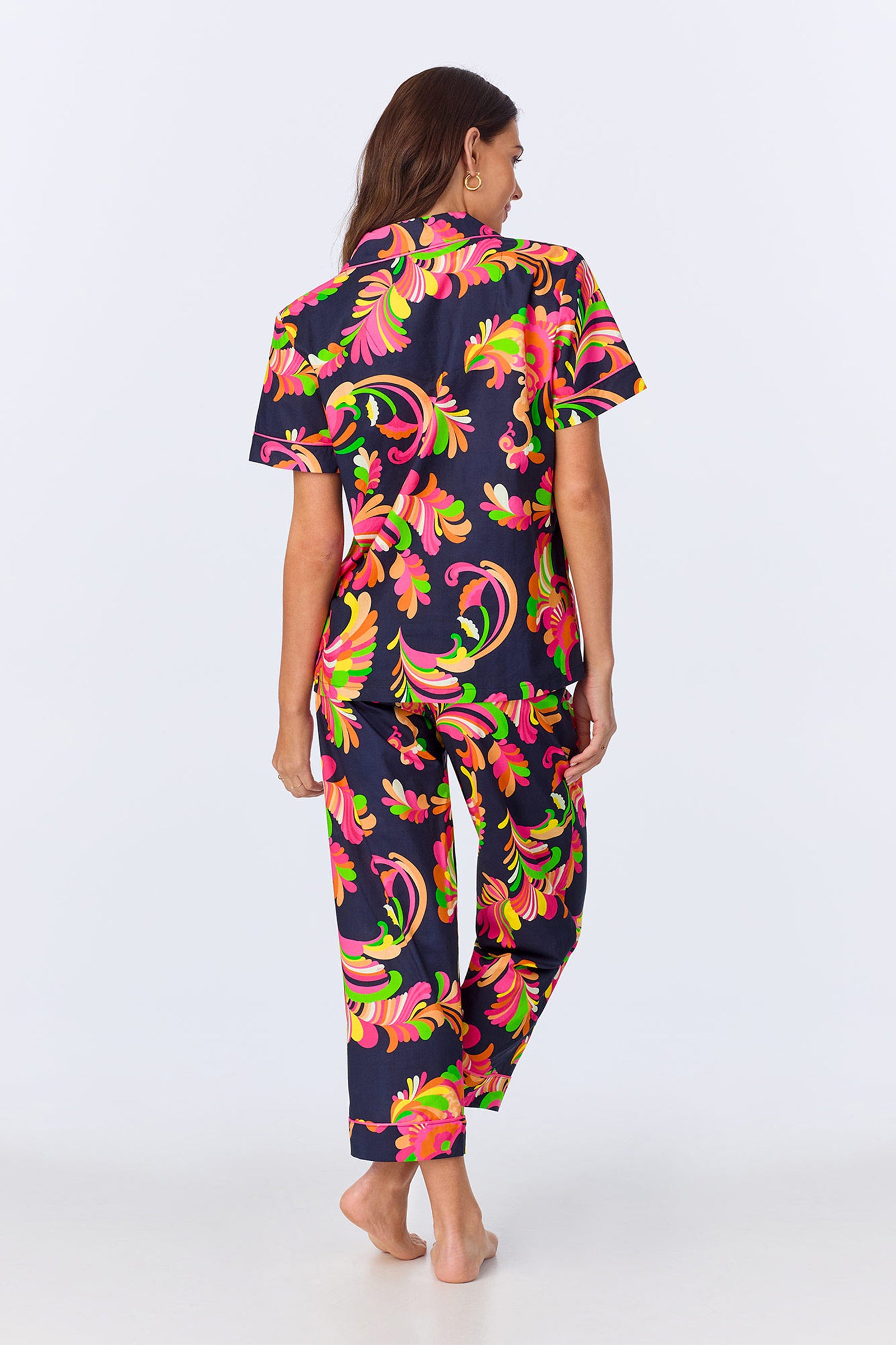 A woman stands barefoot, facing away, wearing the TT SS Crop PJ Set - Theodora Poplin, which features a vibrant abstract floral print in pink, orange, green, and yellow on a dark background.