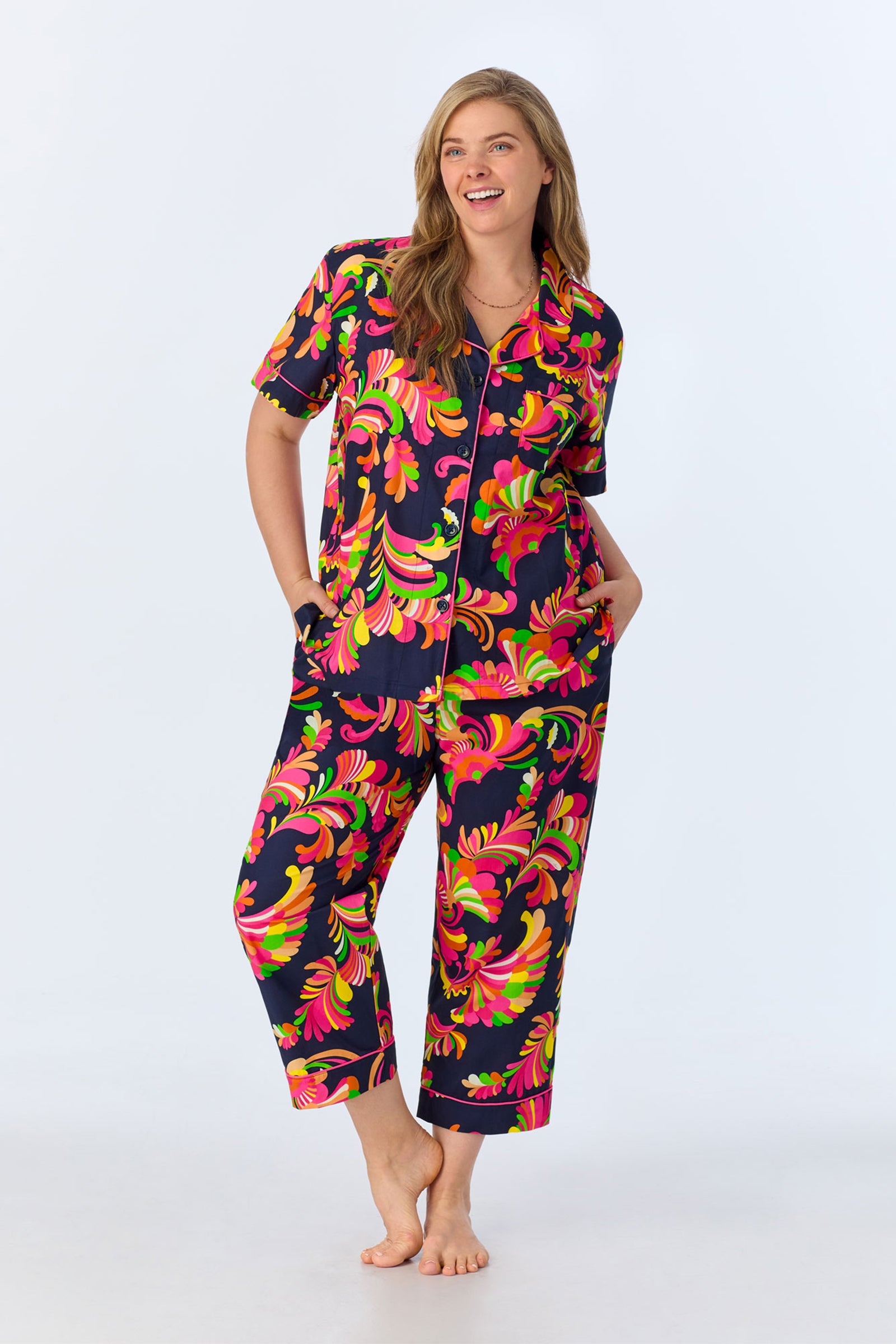 A woman stands barefoot, smiling, wearing the TT SS Crop PJ Set - Theodora Poplin: a matching pajama set featuring a bold floral pattern in pink, green, and yellow on dark poplin with a short-sleeve crop button-up and wide-leg pants.