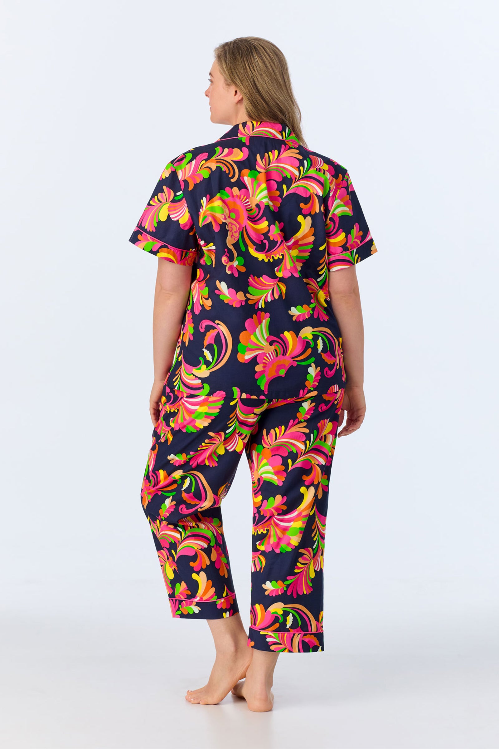 A person stands barefoot facing away, wearing the TT SS Crop PJ Set – Theodora Poplin, which features a matching short-sleeve shirt and pants with a bold, colorful swirling floral pattern on a dark background.
