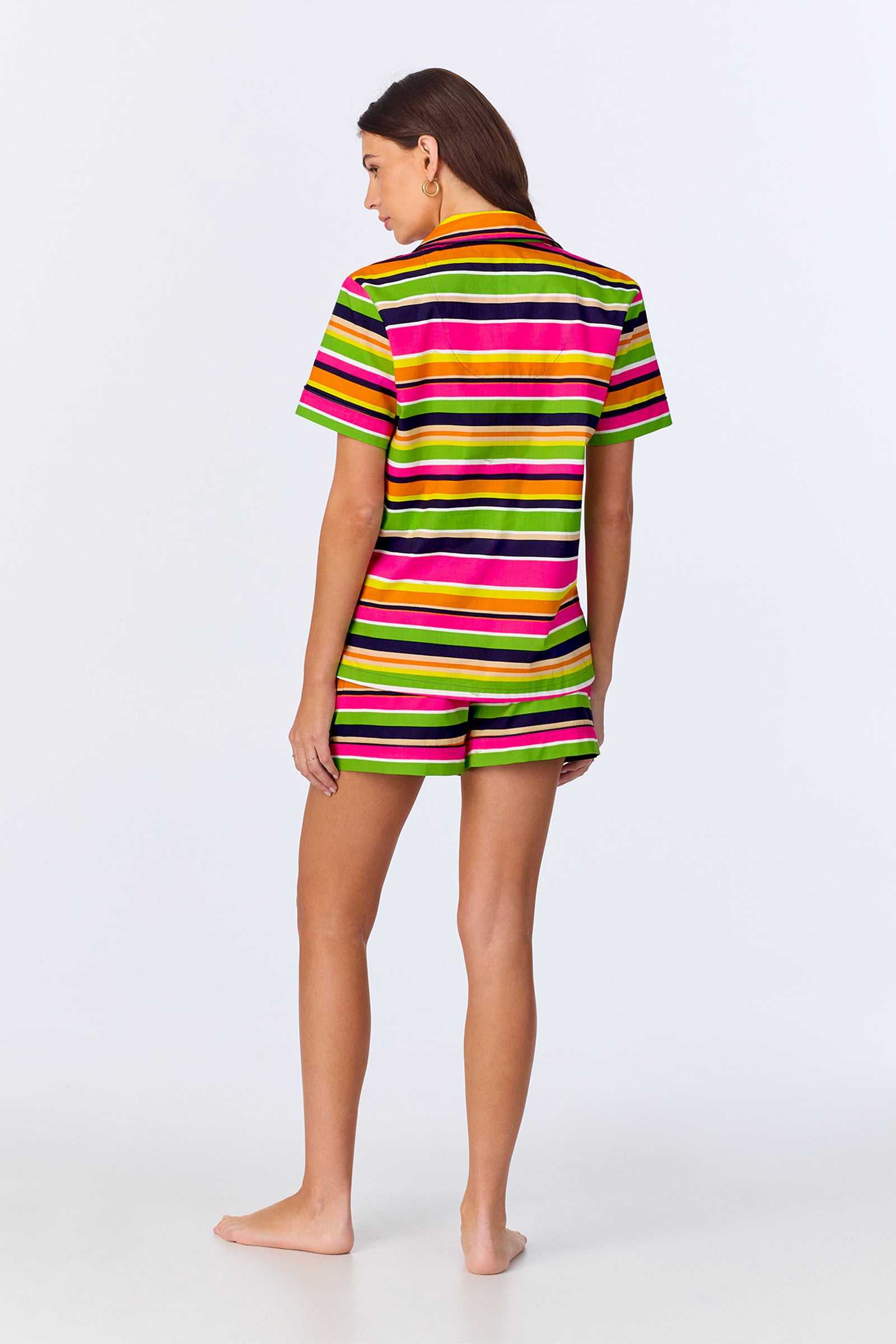 A person stands barefoot, facing away on a plain white background, wearing the TT SS Shorty PJ Set - Poolside Poplin: a matching short-sleeve shirt and shorts with black, yellow, pink, white, and green horizontal stripes.