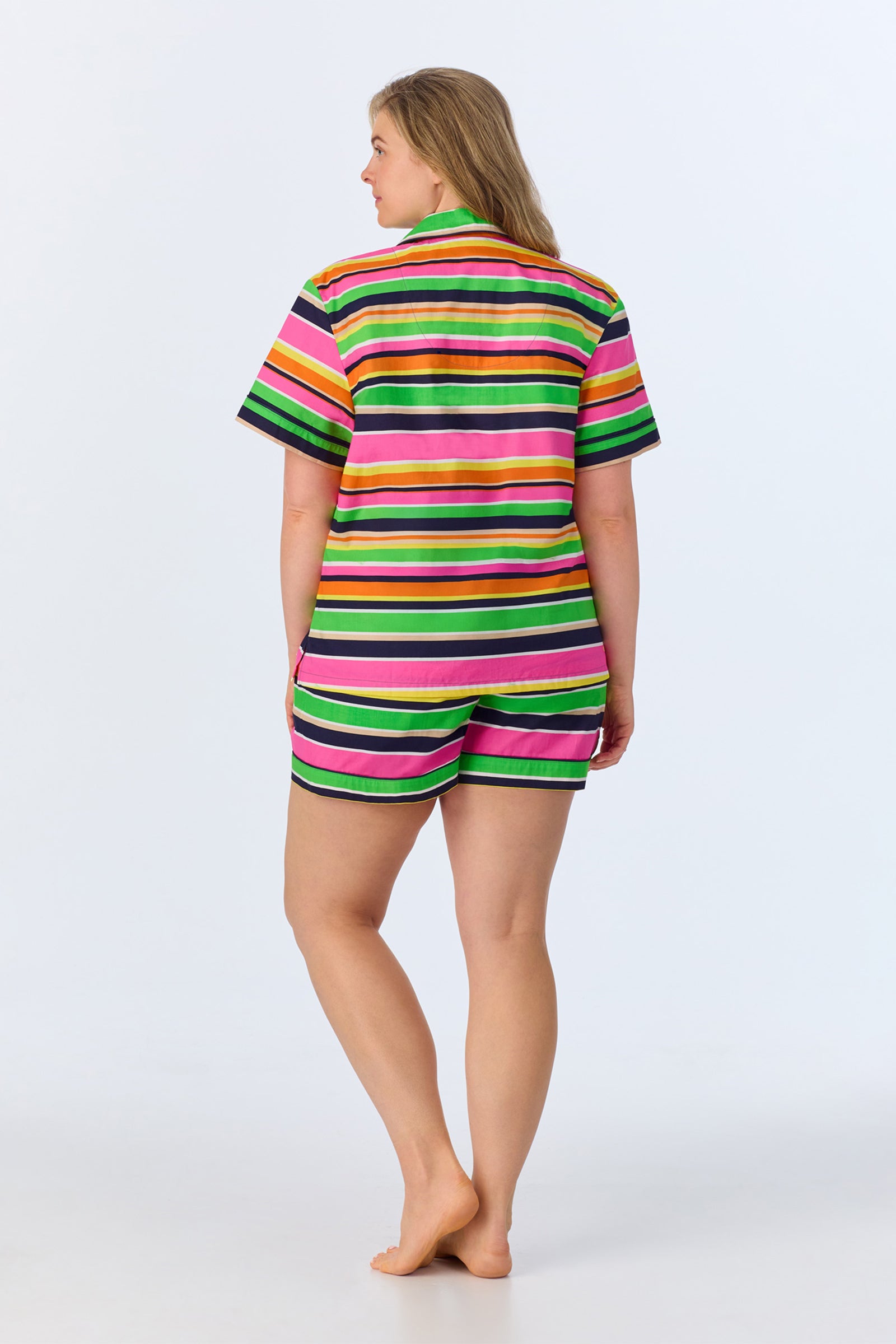 A woman stands barefoot facing away, wearing the TT SS Shorty PJ Set - Poolside Poplin: a matching short-sleeved shirt and shorts with bright pink, green, yellow, and black horizontal stripes on a plain light background.