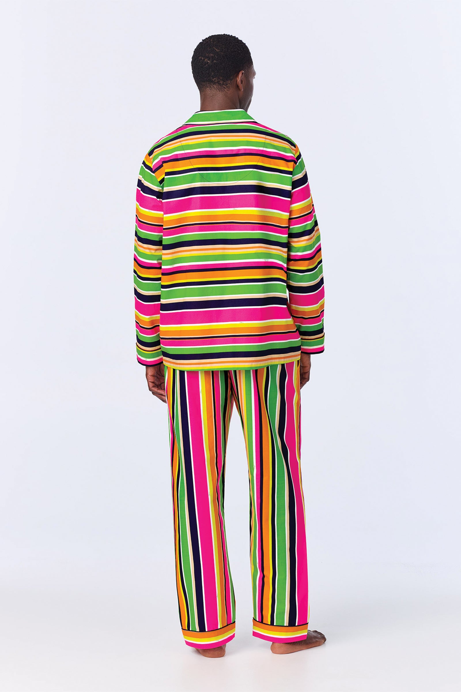 A person stands barefoot facing away, wearing the MT LS Pant PJ Set - Poolside Stripe Poplin, featuring bold horizontal and vertical stripes in pink, green, yellow, white, and black on a plain light background.