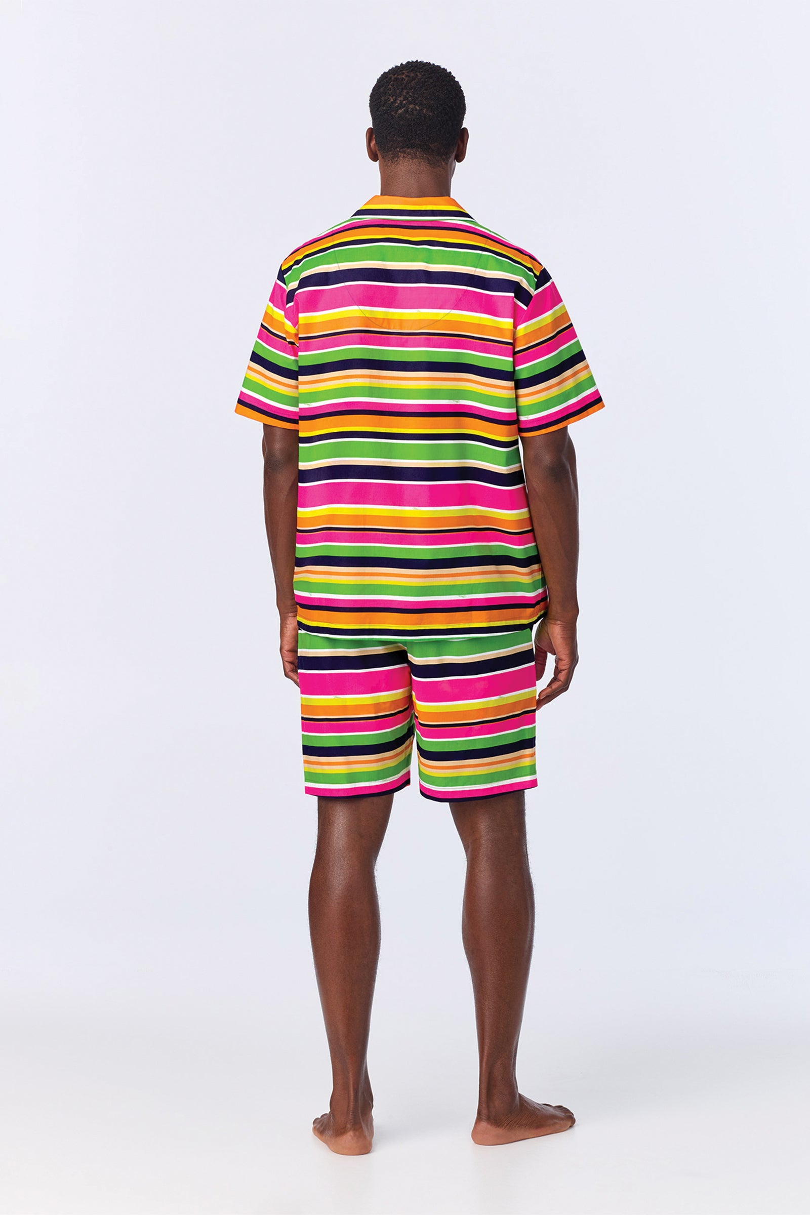 A person stands barefoot, facing away, wearing the MT SS Boxer PJ Set - Poolside Stripe Poplin featuring bold horizontal stripes in yellow, green, pink, black, and white against a plain light background.
