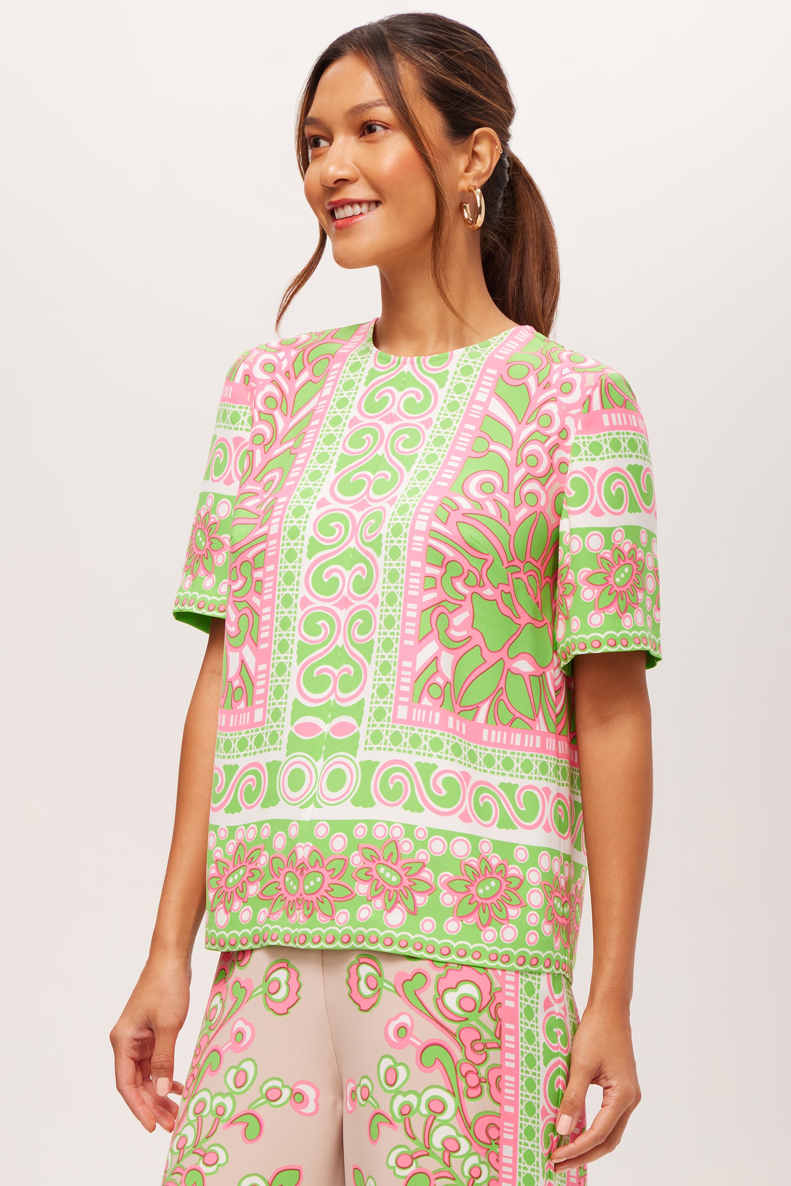 A woman with long brown hair tied back smiles and faces slightly to the side, wearing the Riggs Short-Sleeve Top in Petra Floral against a plain white background.