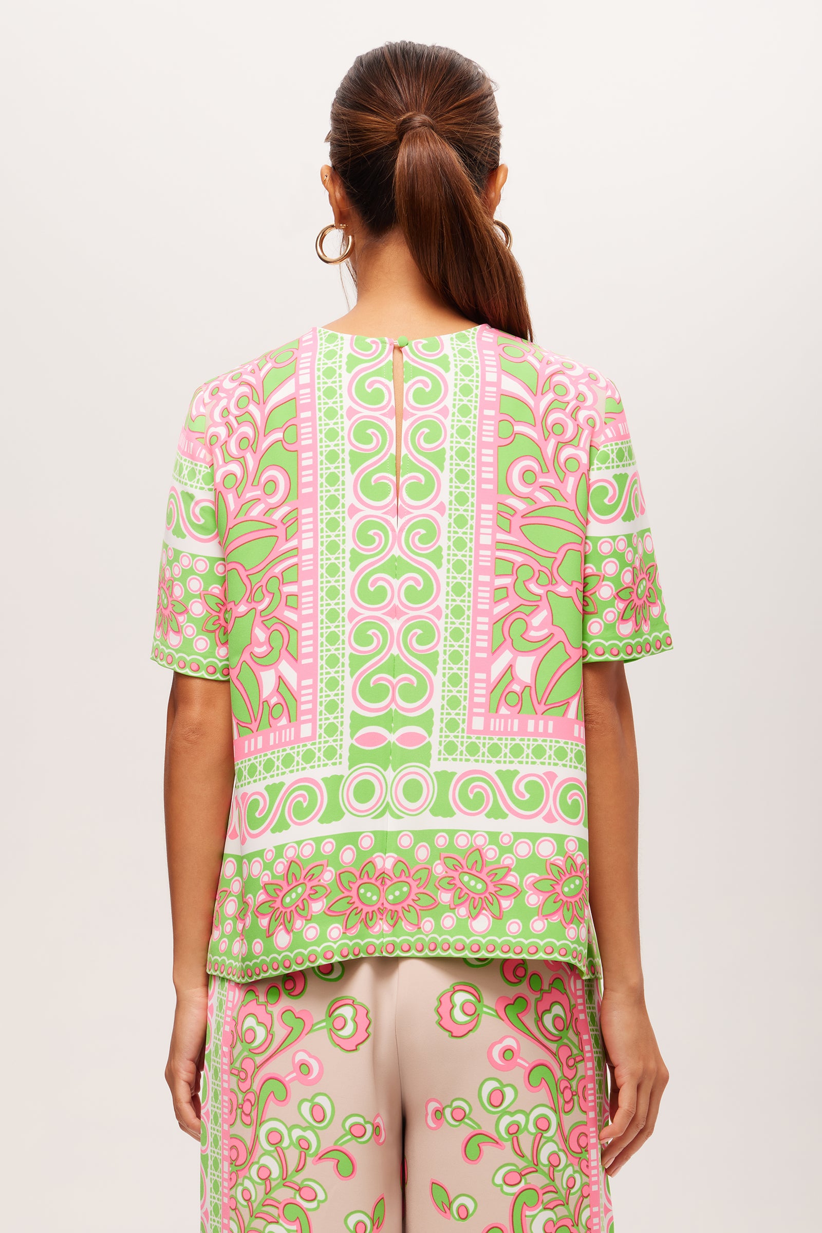 A woman with long brown hair in a low ponytail is seen from behind against a plain background, wearing the Riggs Short-Sleeve Top - Petra Floral, which features pink, green, and cream geometric and floral patterns.