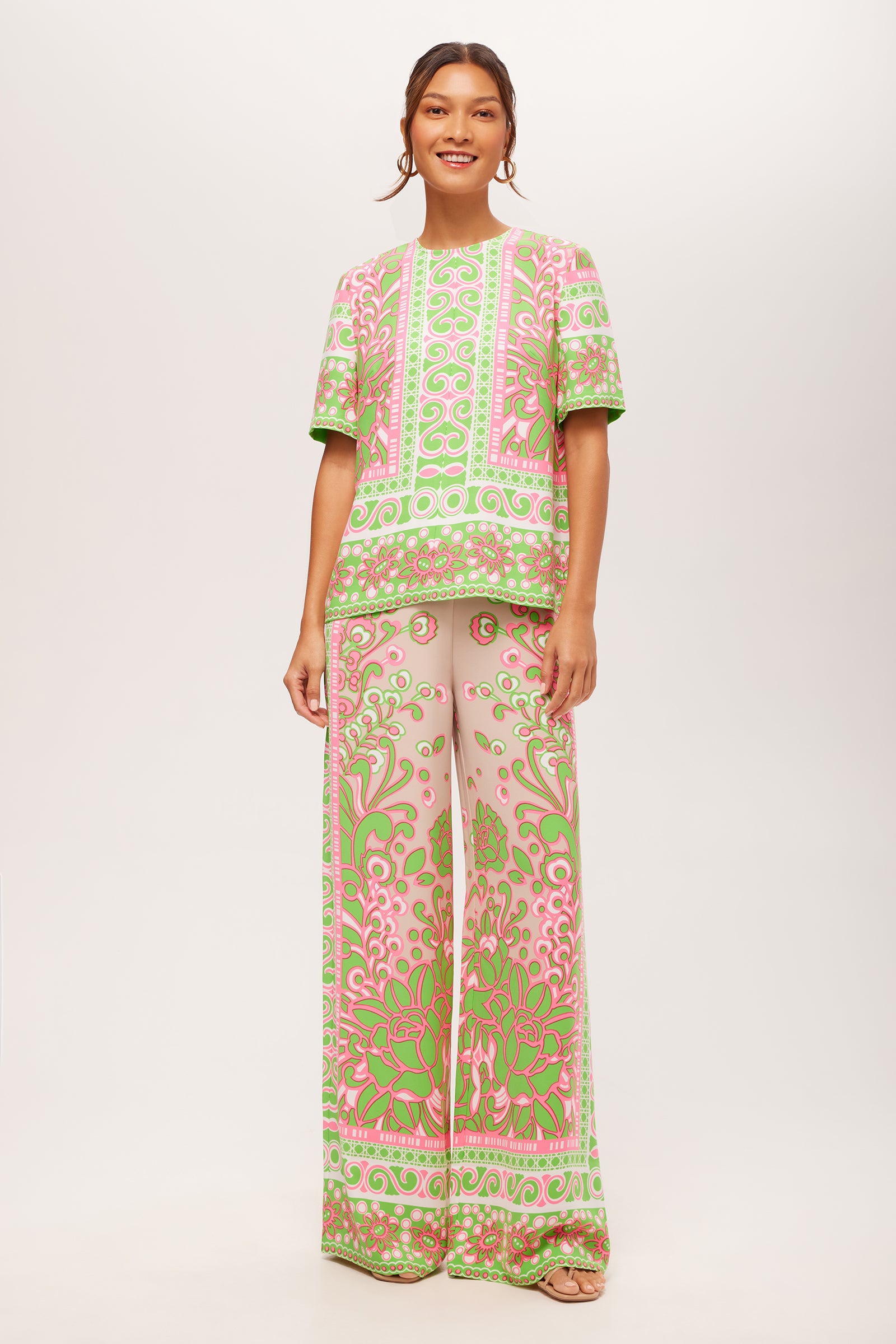 A woman smiles against a plain light background, wearing the Federer Wide-Leg Pant in Petra Floral—a matching set featuring an ornate pink, green, and beige swirling floral pattern with a short-sleeve top and wide-leg pants. Her hair is tied back.