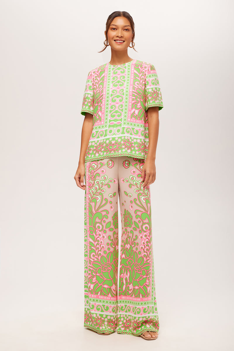 A woman smiles against a plain light background, wearing the Federer Wide-Leg Pant in Petra Floral—a matching set featuring an ornate pink, green, and beige swirling floral pattern with a short-sleeve top and wide-leg pants. Her hair is tied back.