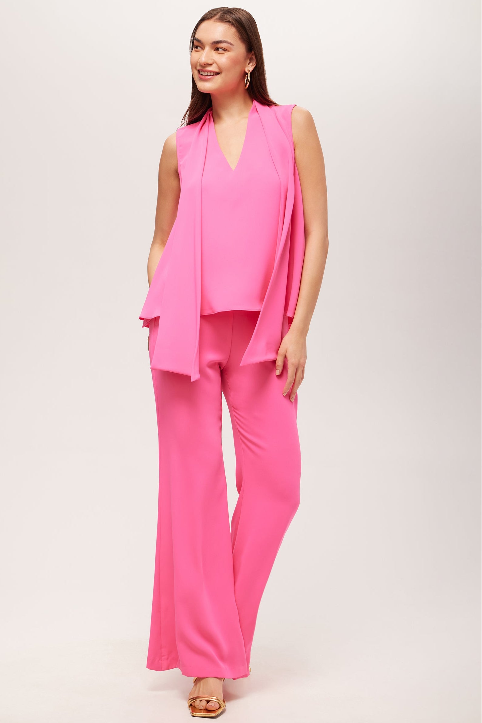 A woman smiles in the Circuit Sleeveless V-Neck Top - Double Georgette, a bright pink sleeveless v-neck with matching scarf detail, styled with wide-leg pants and beige heels against a light background.