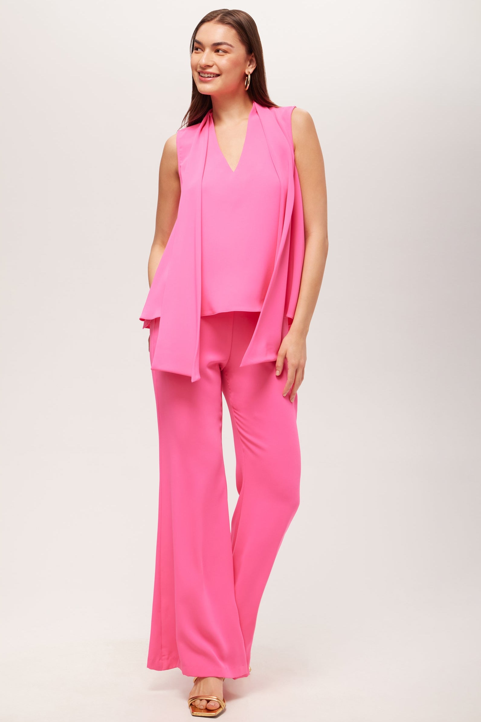 A woman smiles, wearing the Drop Shot Flared Pant - Double Georgette in bright pink with a matching V-neck sleeveless top, scarf, hoop earrings, and open-toe sandals. She has long brown hair and stands against a light background.