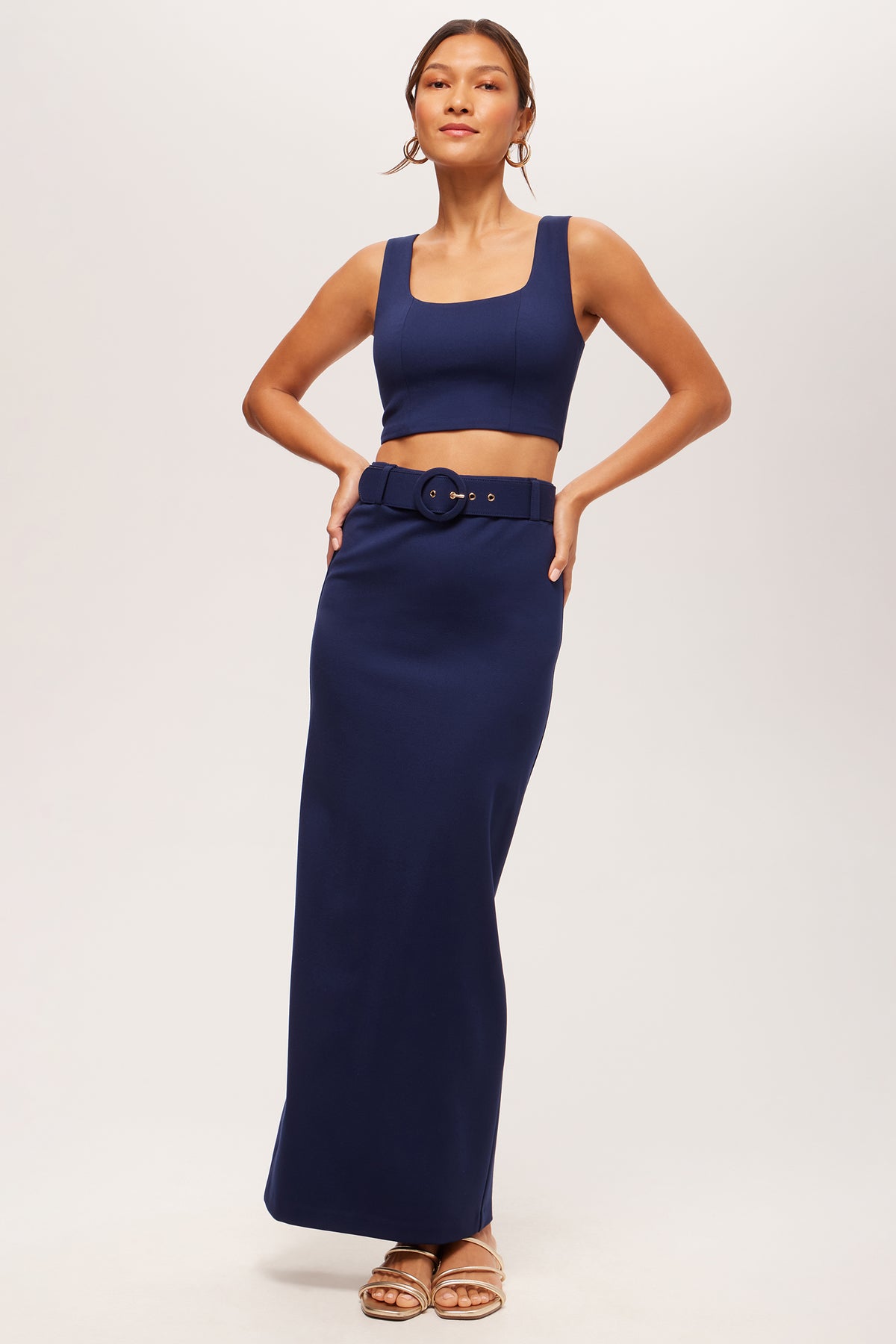 A woman models the Grand Slam Sleeveless Crop Top - Cozy Ponte in navy blue, paired with a matching high-waisted skirt, large belt, sandals, and hoop earrings against a plain light background.