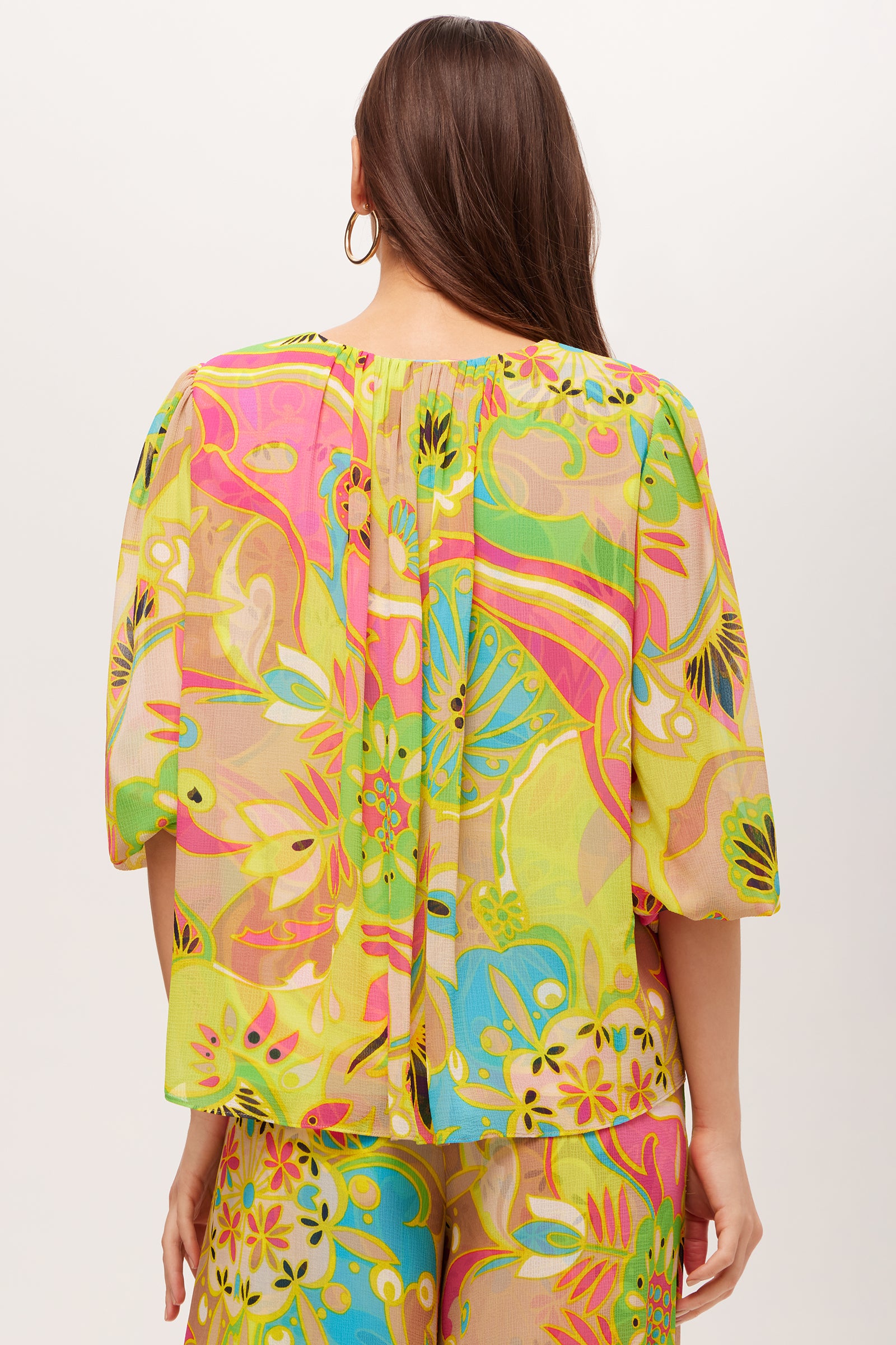 A woman with long brown hair is seen from behind wearing the Sampras 3/4-Sleeve Top - Venus, a colorful floral blouse in vibrant pink, yellow, green, and blue tones, paired with matching pants.