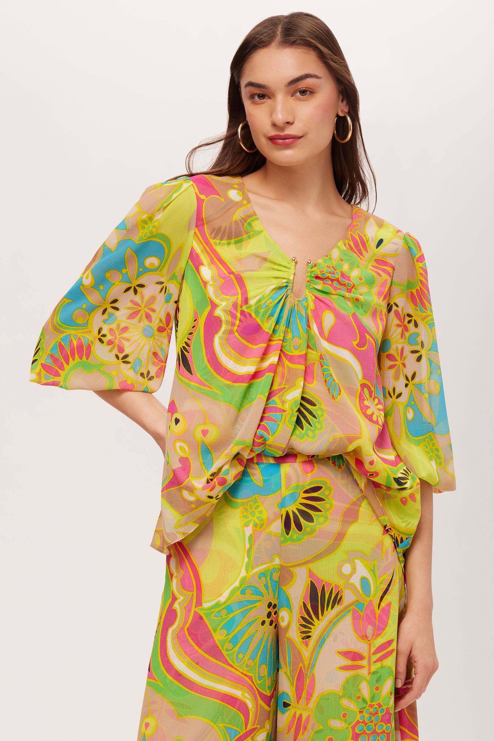 A woman wears the Sampras 3/4-Sleeve Top in Venus, featuring vibrant swirling designs in yellow, green, pink, and blue, paired with matching pants against a plain white background.