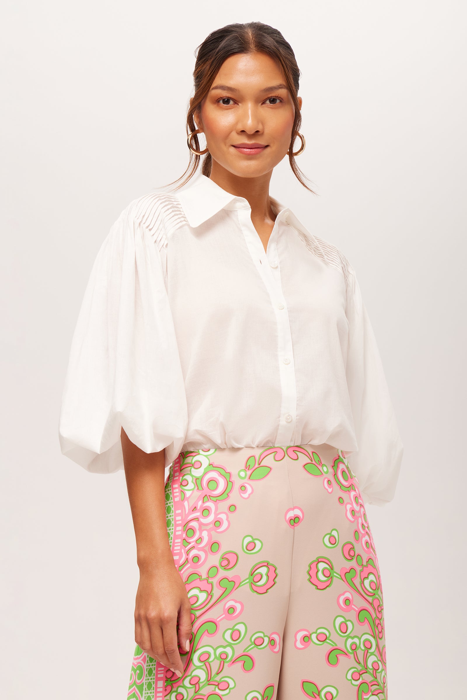 A woman in the white Court Short-Sleeve Button-Up Top with voluminous sleeves and high-waisted, light pink floral pants stands against a plain white background.